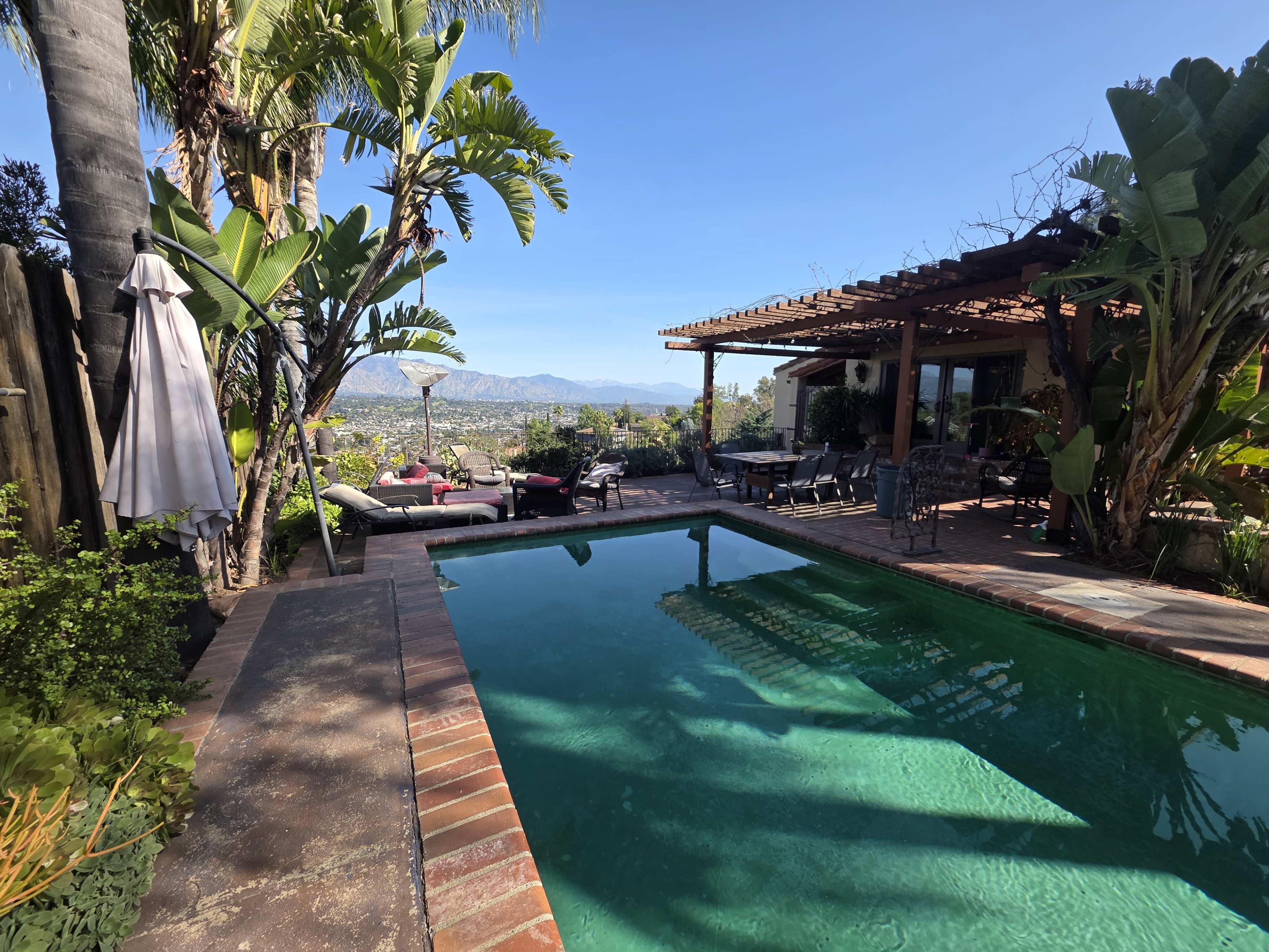 A swimming pool reflects the surrounding greenery and mountains, with outdoor seating and a pergola visible nearby.