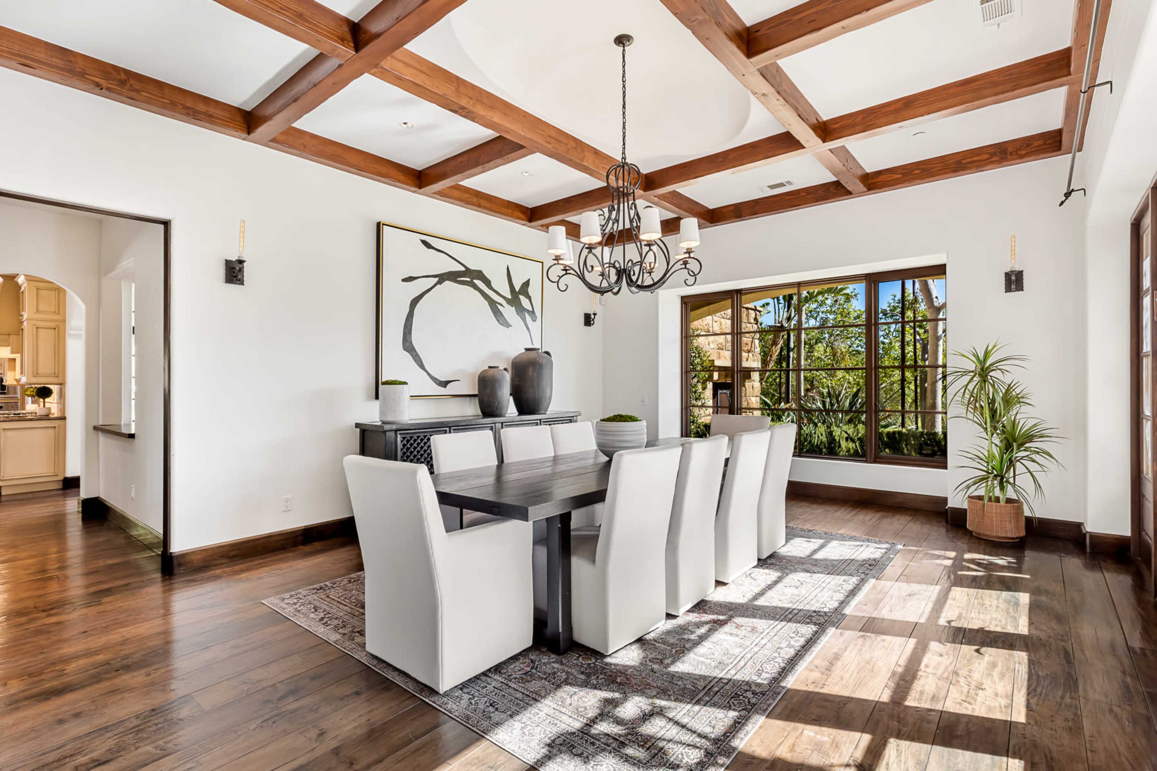 A modern dining room features a large rectangular table surrounded by six upholstered chairs, with a chandelier overhead and a large window providing natural light.