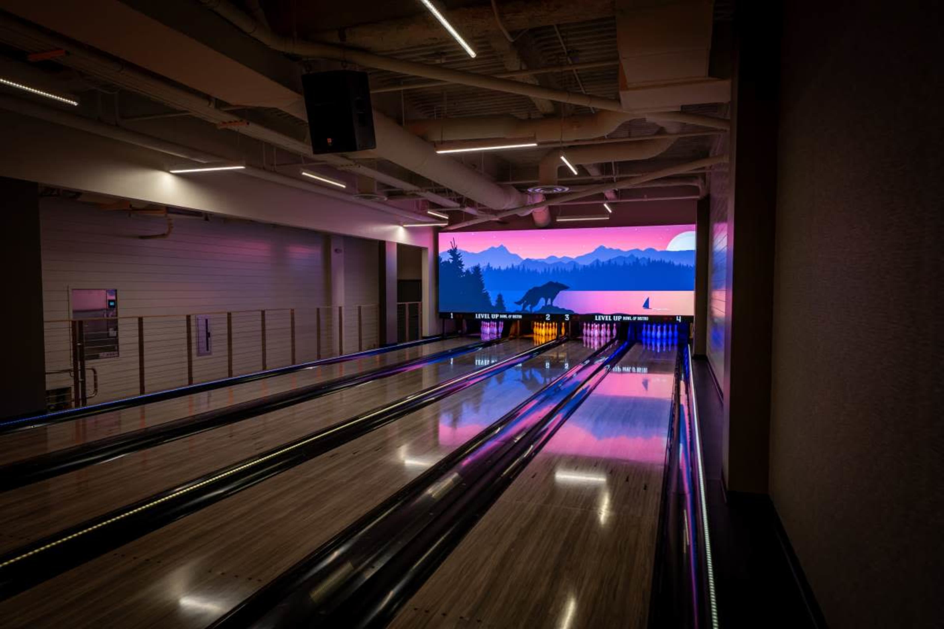 The image shows a modern bowling alley with polished wooden lanes and a large, illuminated mural of a mountain landscape in the background.