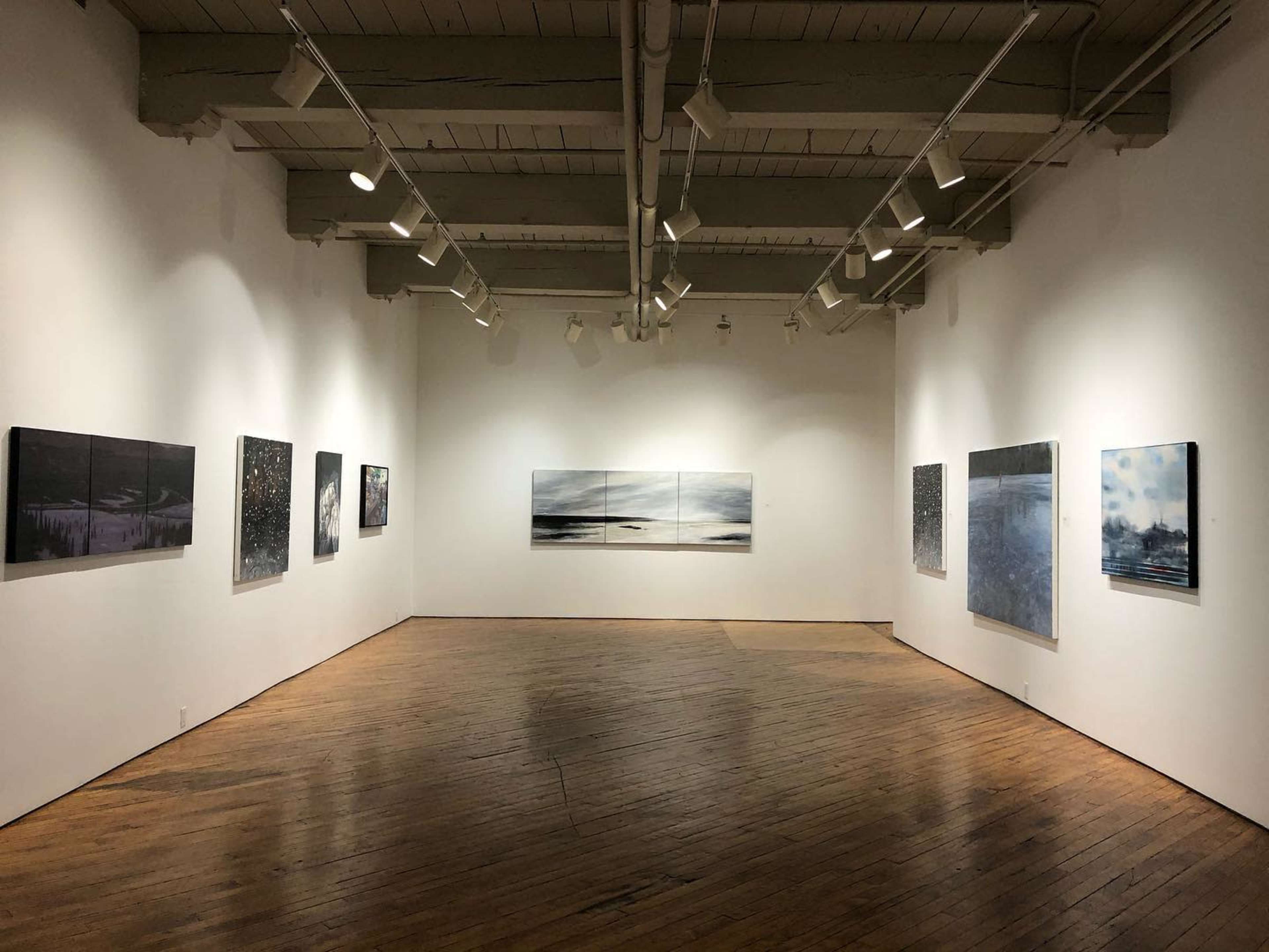 A spacious art gallery with wooden floors and white walls, featuring several framed artworks hung at eye level.