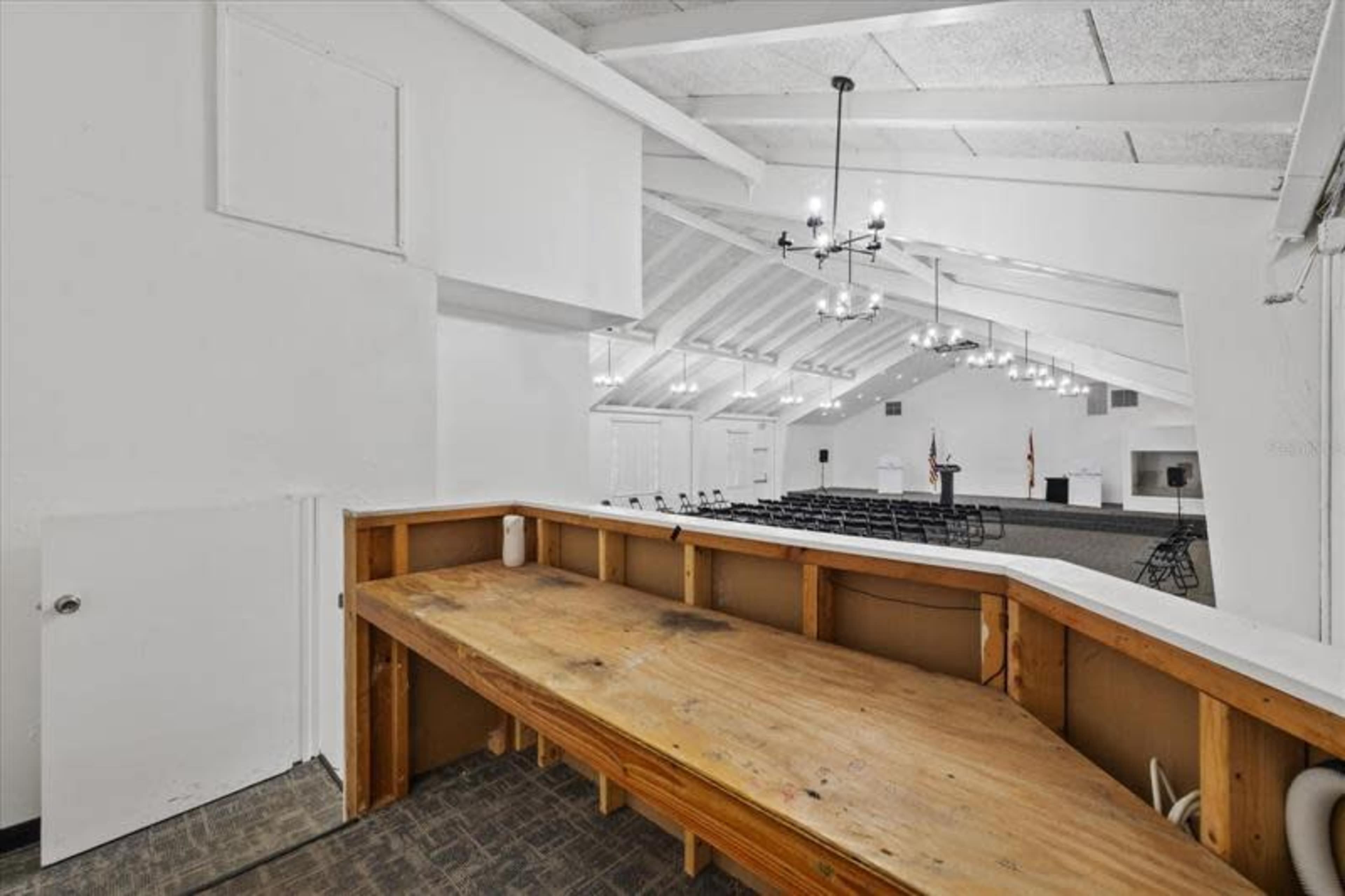 The image shows a spacious, brightly lit room with vaulted ceilings, featuring a stage at the far end and rows of chairs set up for an event.