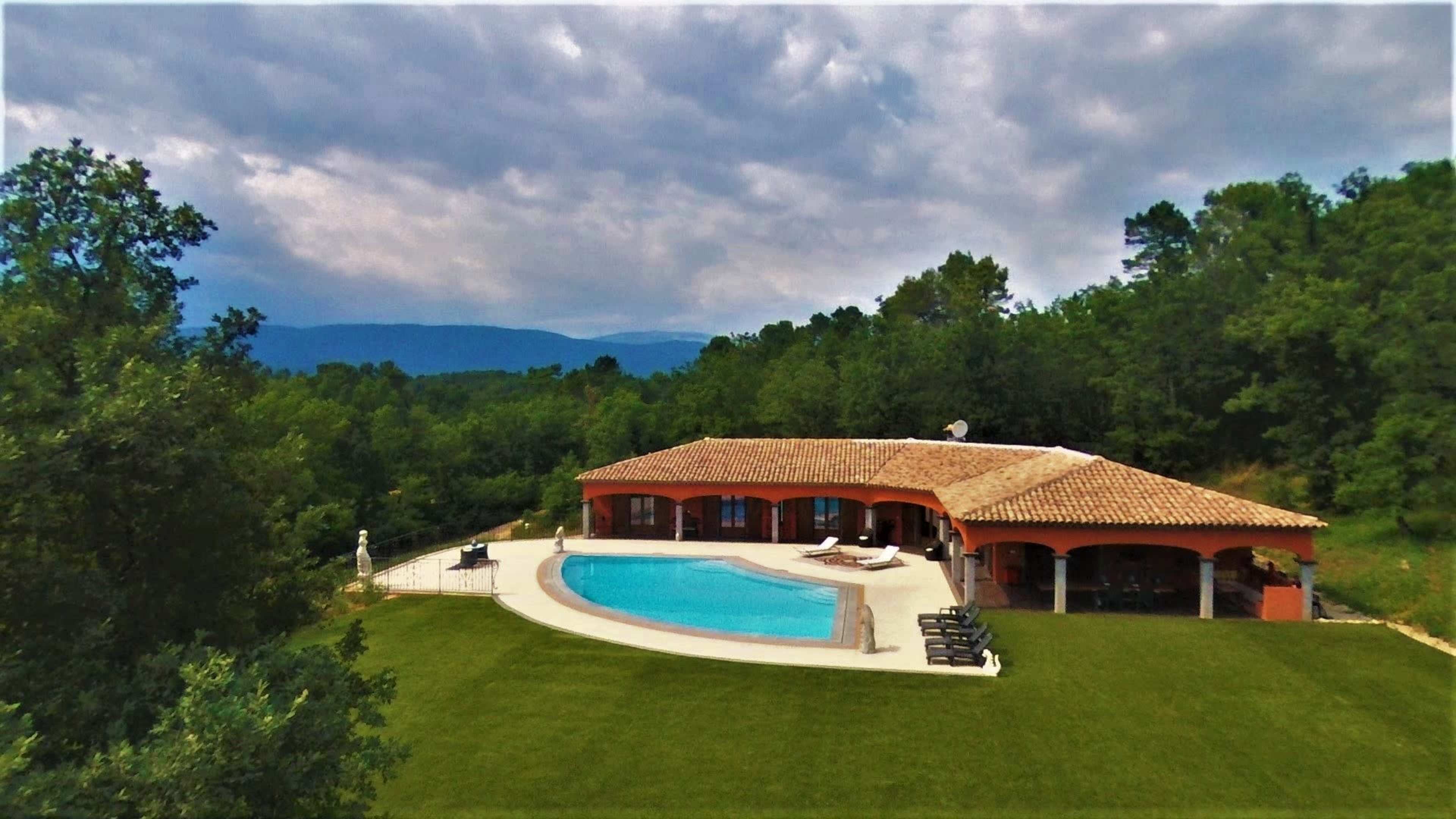 The image shows a modern villa with a swimming pool, set in a green landscape surrounded by trees and mountains in the background.