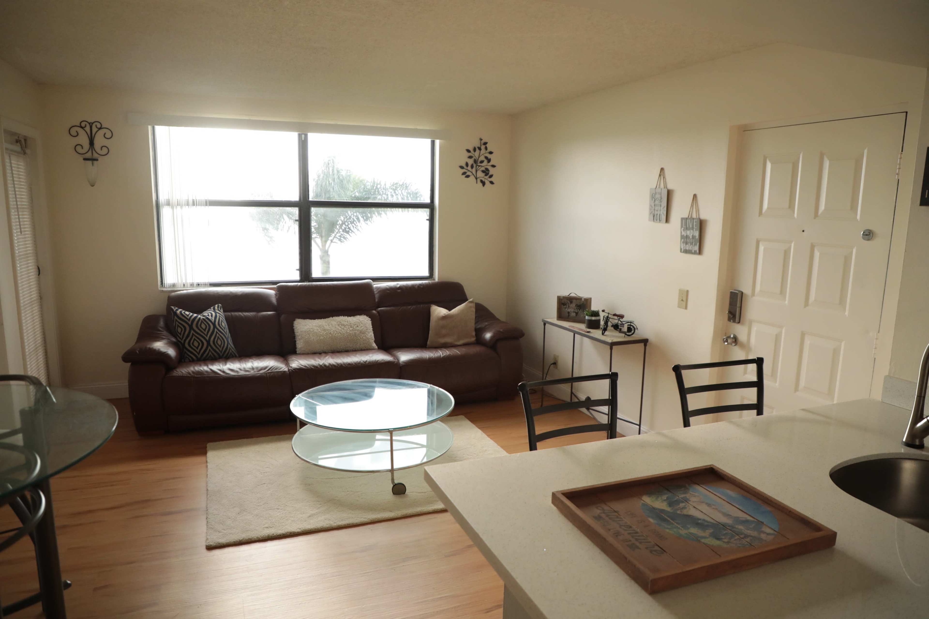 A living room with a brown leather couch, a glass coffee table, and a large window allowing natural light to enter the space.