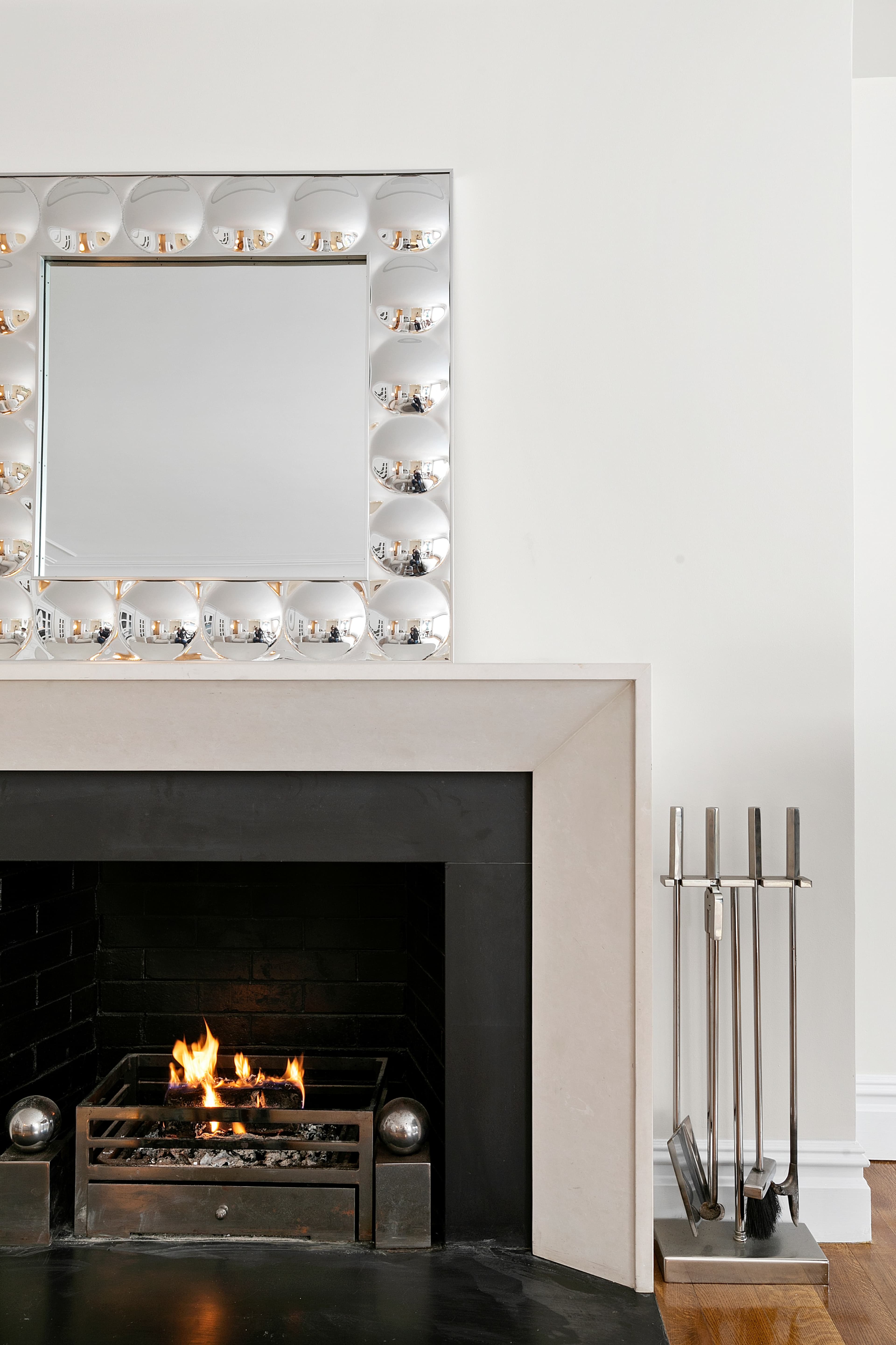 A modern fireplace with an open flame, a minimalist mirrored frame above it, and a set of sleek tools beside the hearth.