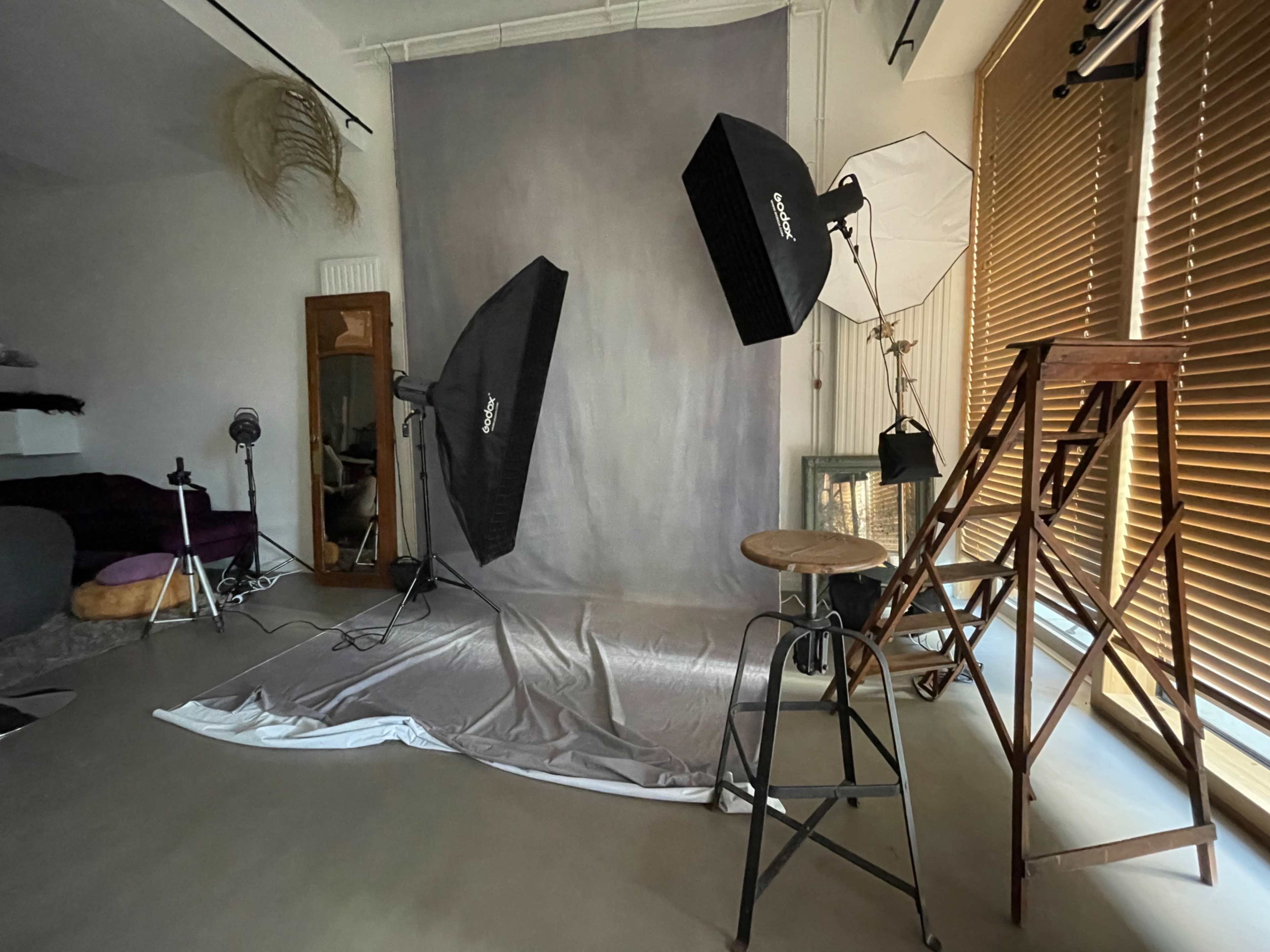 A photography studio setup with two softbox lights, a gray backdrop, a wooden ladder, and various equipment placed around the room.
