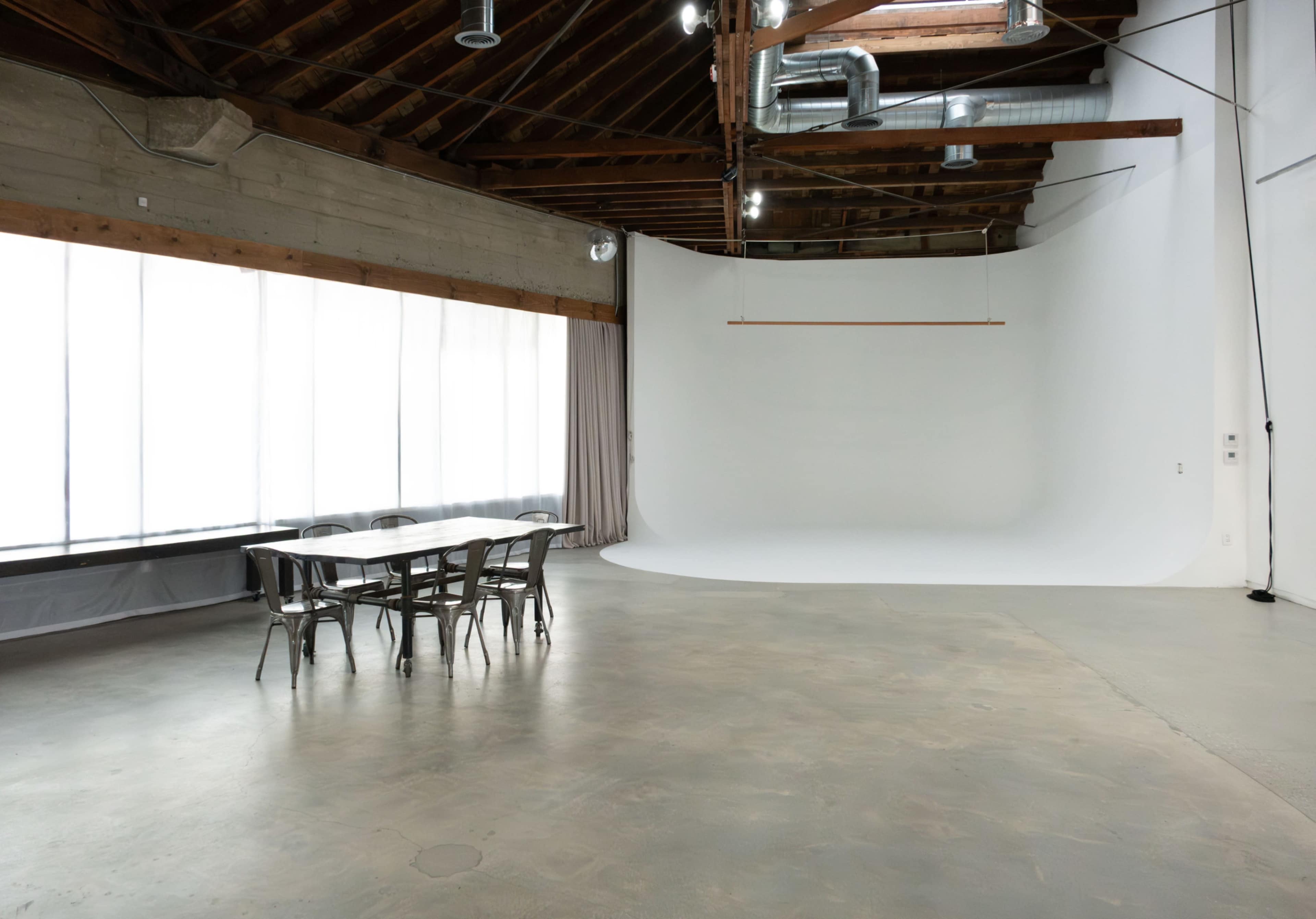 The image shows a spacious, well-lit studio with a large blank backdrop, a wooden ceiling, and a metal table surrounded by chairs.