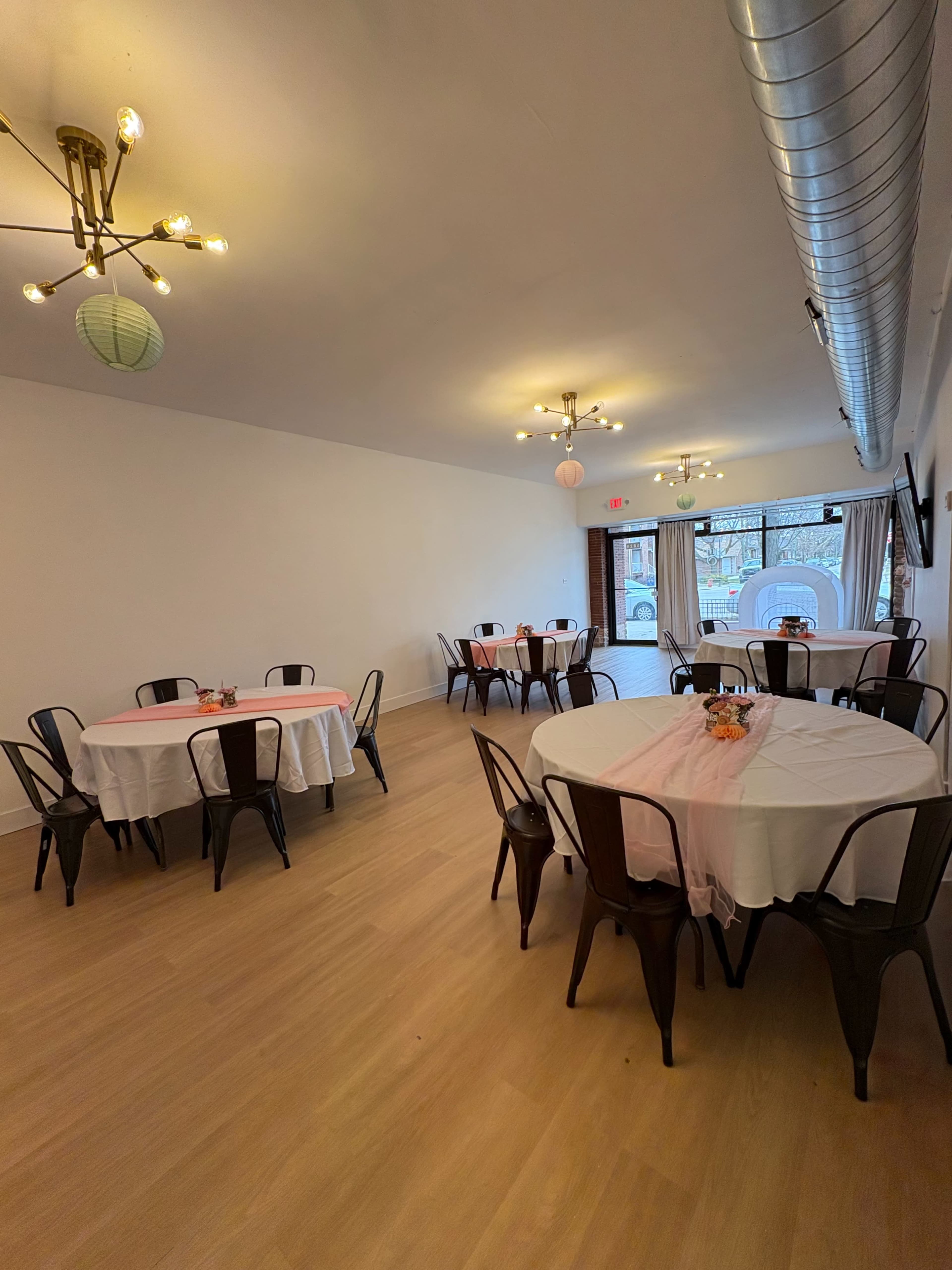 The image shows a spacious event room with several round tables covered in white tablecloths, each adorned with a small centerpiece, and equipped with black metal chairs.