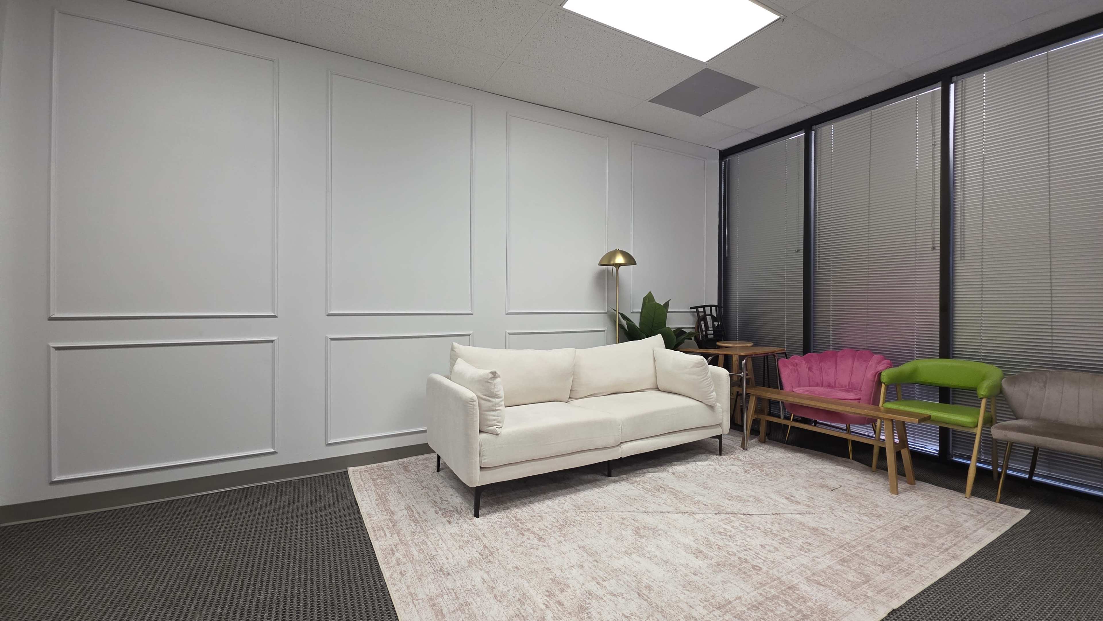 The image shows a small, minimalist room with a white couch, a light-colored rug, and a few pieces of furniture against a backdrop of paneled walls and large windows.