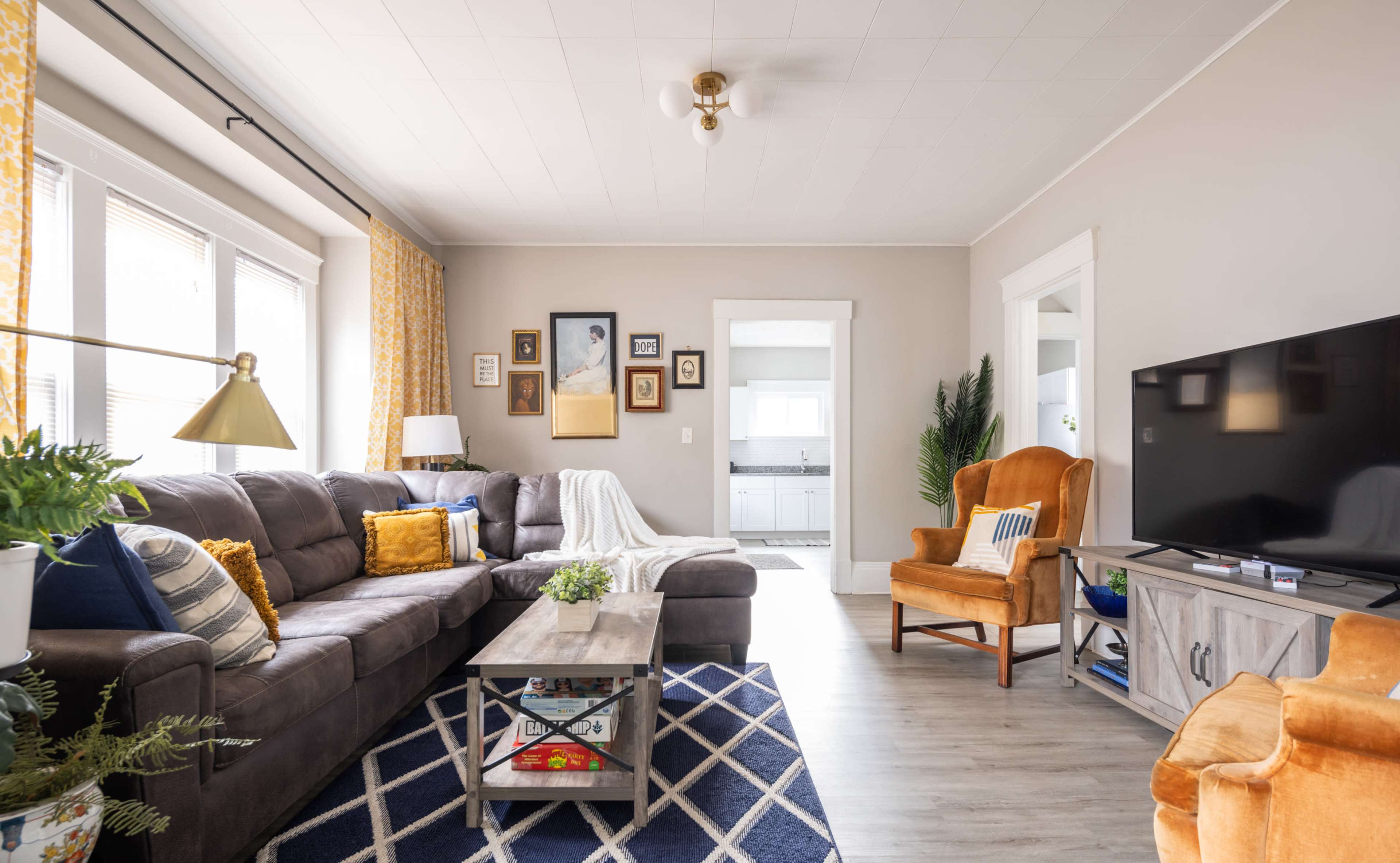 The living room features a gray sectional couch, a wooden coffee table, an orange armchair, and a television against a light-colored wall, with decorative curtains and framed pictures.