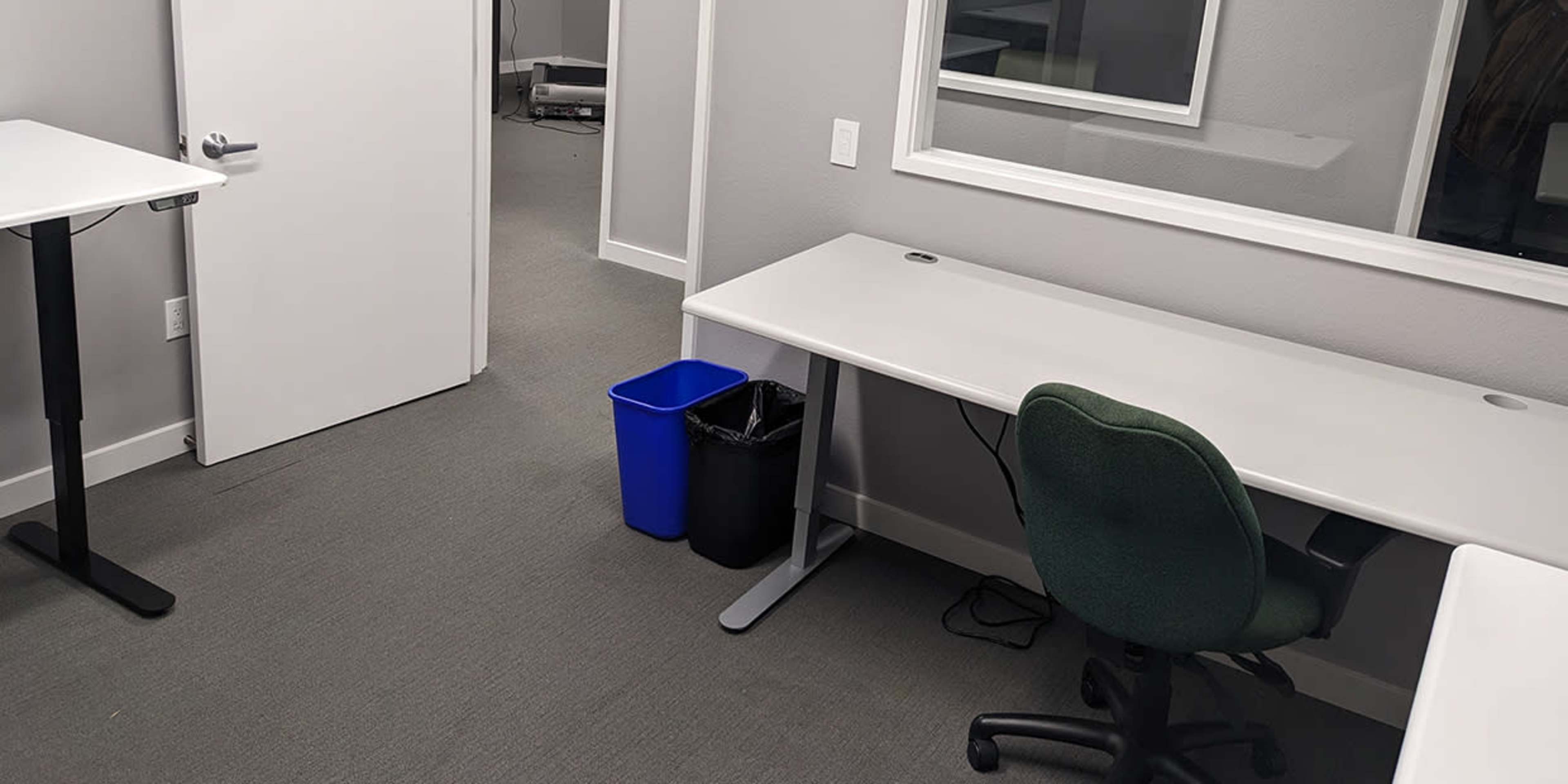 A minimalist office space features two desks, a blue recycling bin, and a green office chair, with a door and a mirrored wall visible.