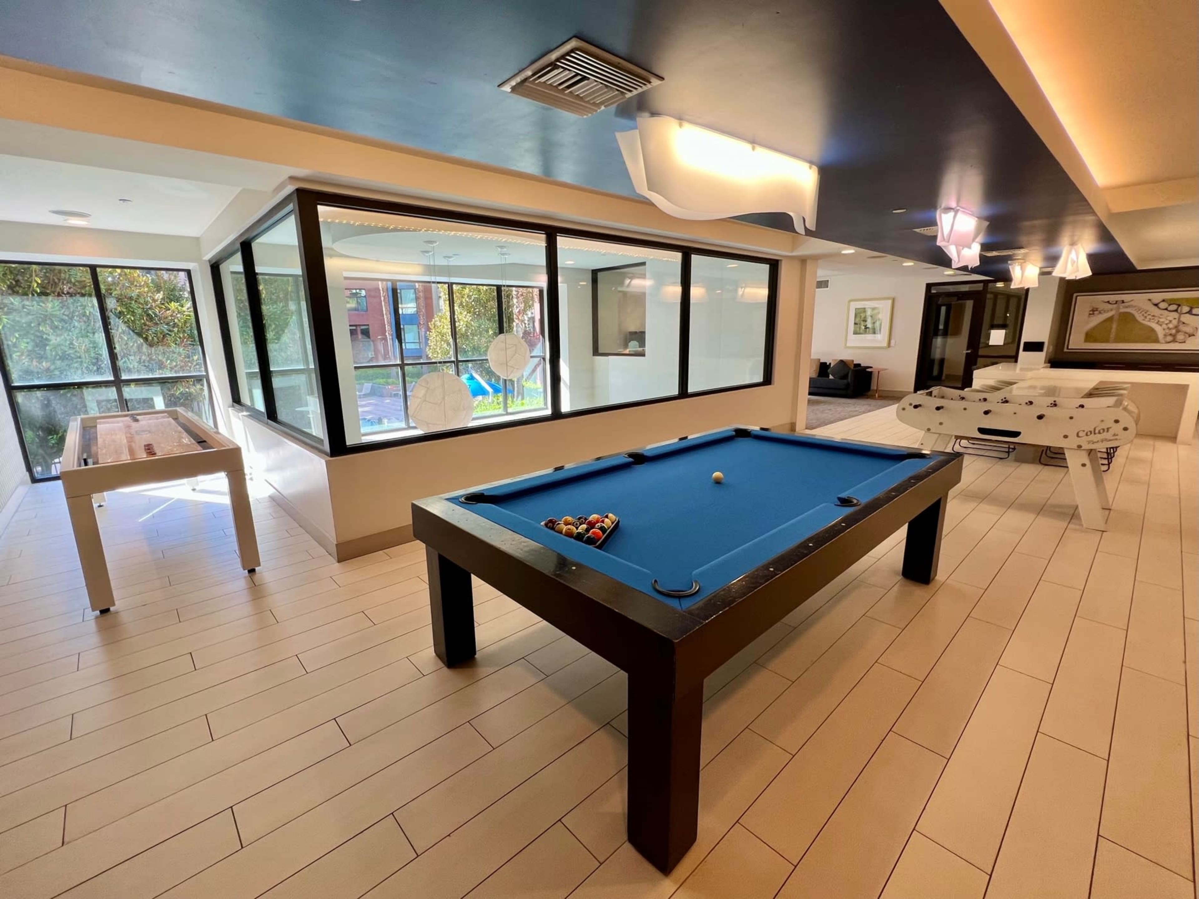 A recreational room featuring a blue billiards table, a foosball table, and large windows with outdoor views.