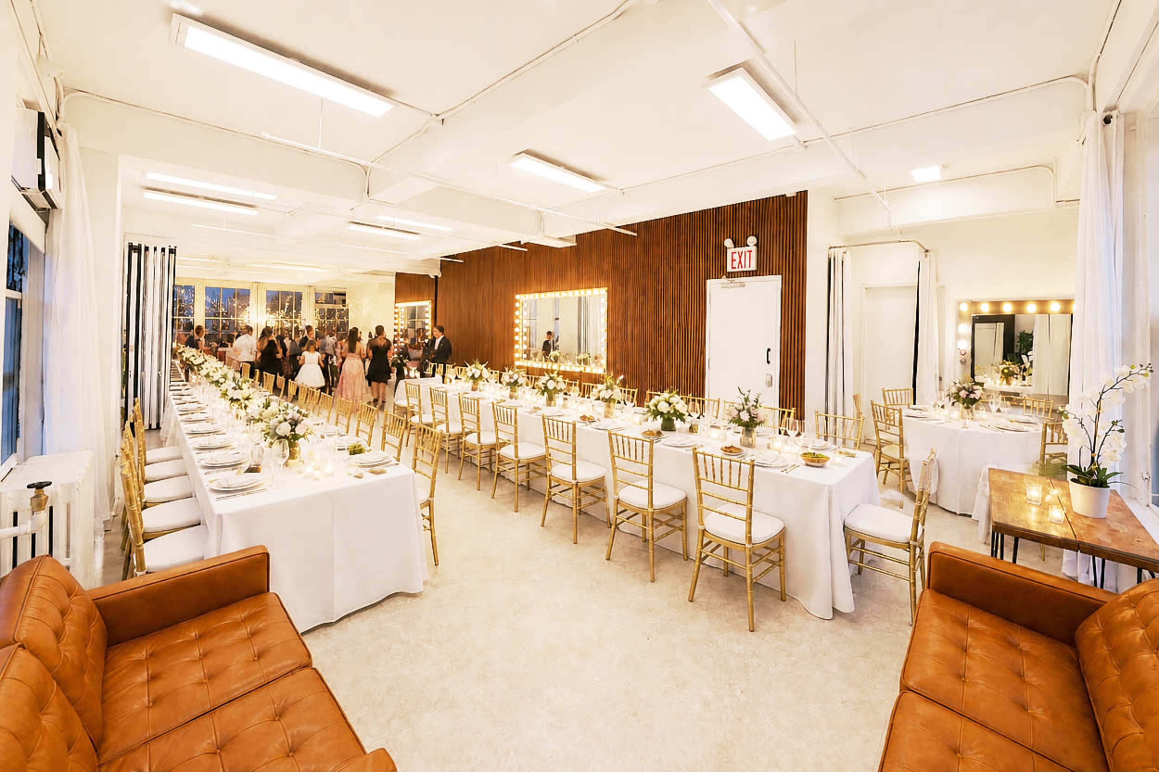 The image shows a spacious banquet hall set with multiple long tables adorned with white tablecloths and gold chairs, while a gathering of people can be seen in the background.