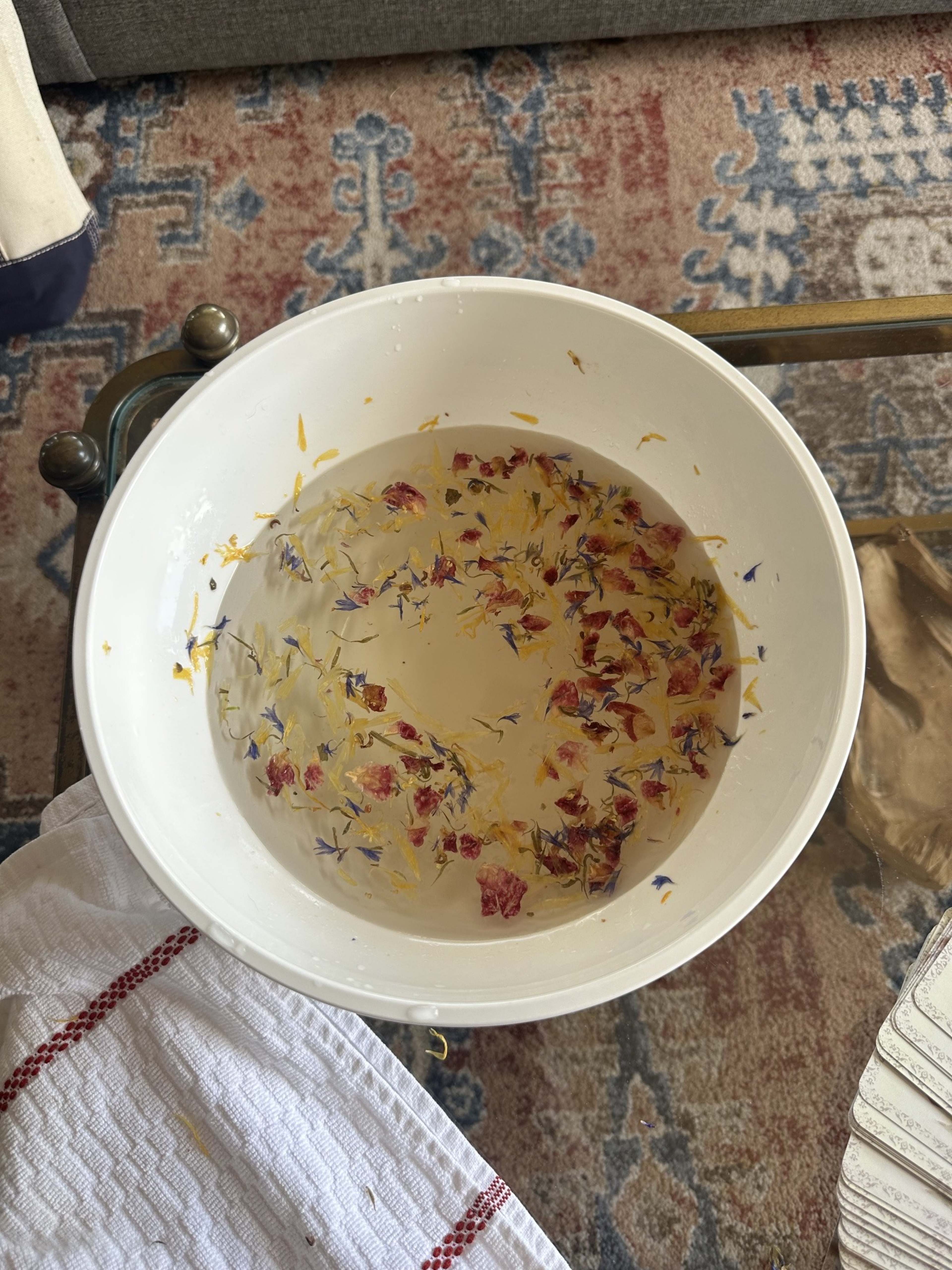 A white bowl filled with water and floating dried flowers sits on a table next to a folded cloth.
