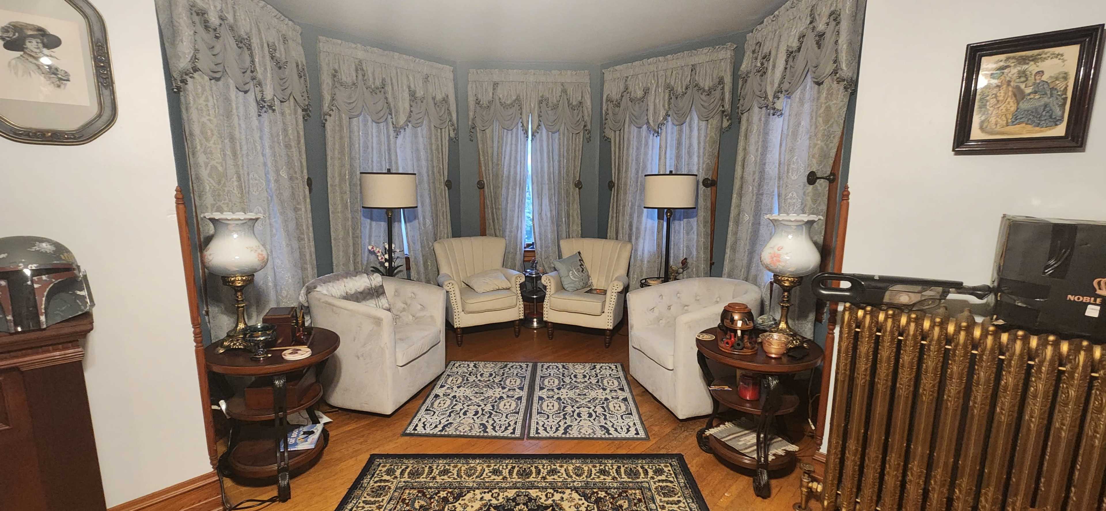 The image shows a cozy living room with two upholstered chairs facing each other, surrounded by decorative lamps, a patterned rug, and ornate window treatments.