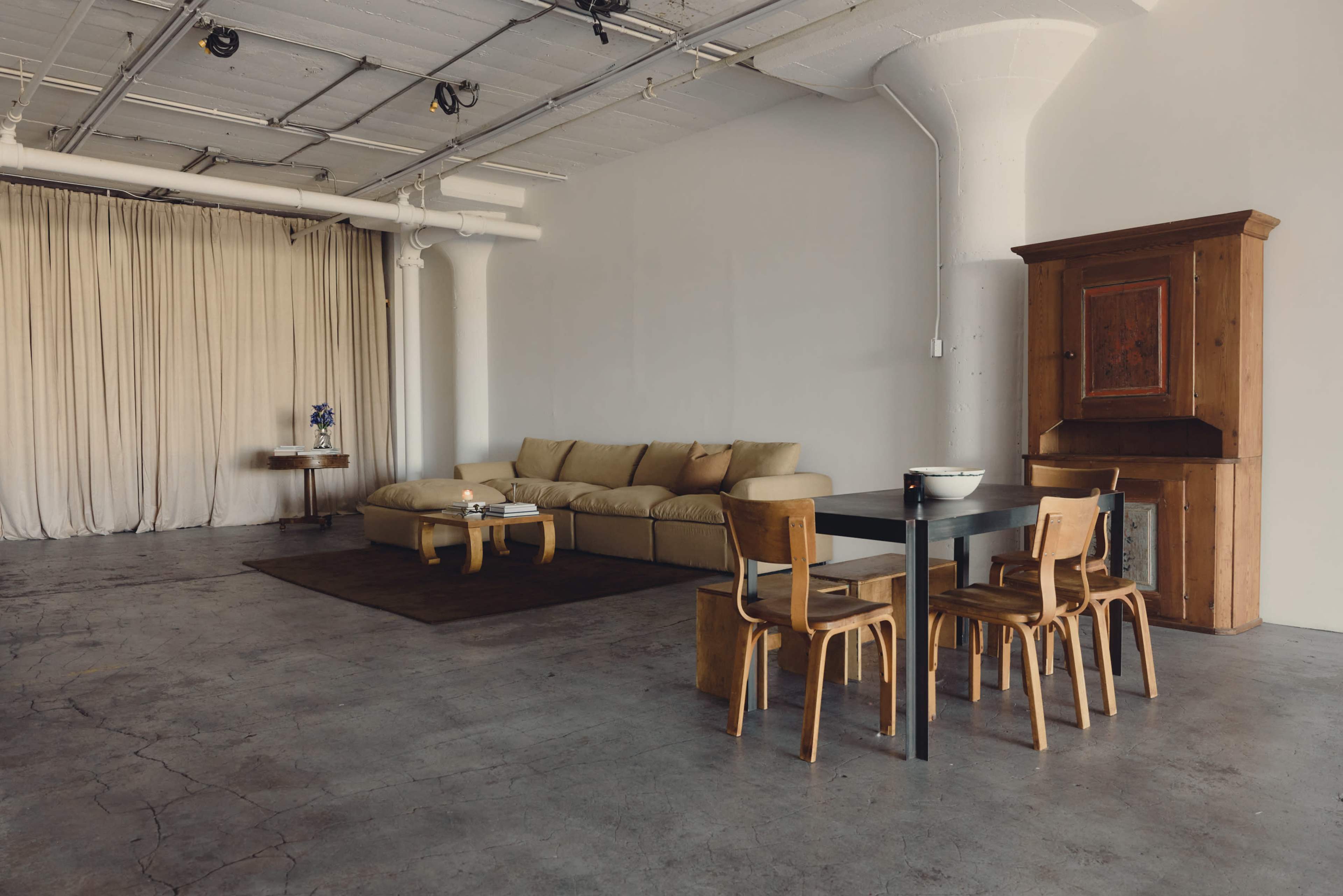 The image shows a spacious living area with a large beige sofa, a wooden dining table with four chairs, and a tall cabinet, all set against a backdrop of light gray walls and curtains.