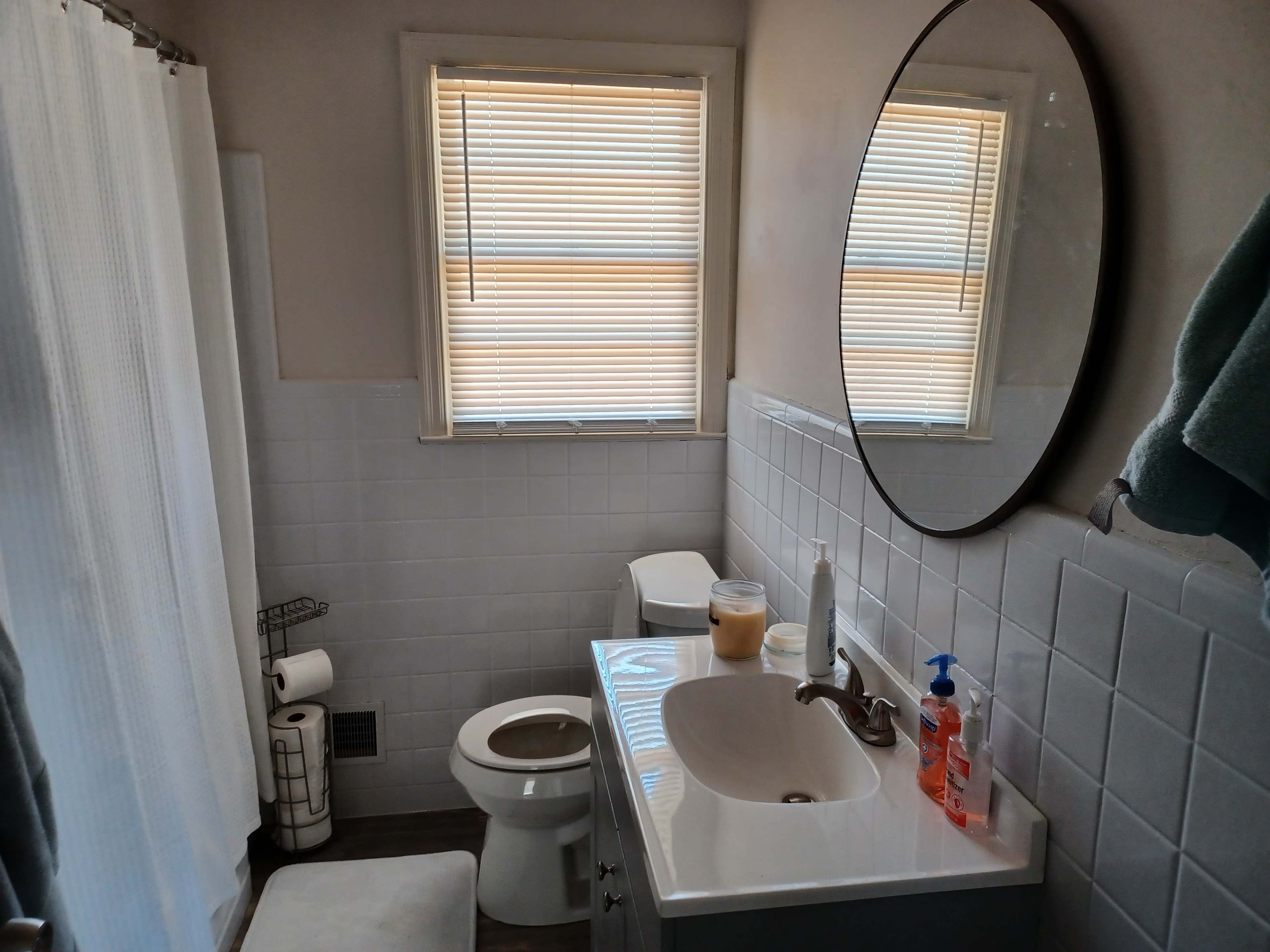 A bathroom features a round mirror above a sink, a toilet, a shower curtain, and a window with blinds.