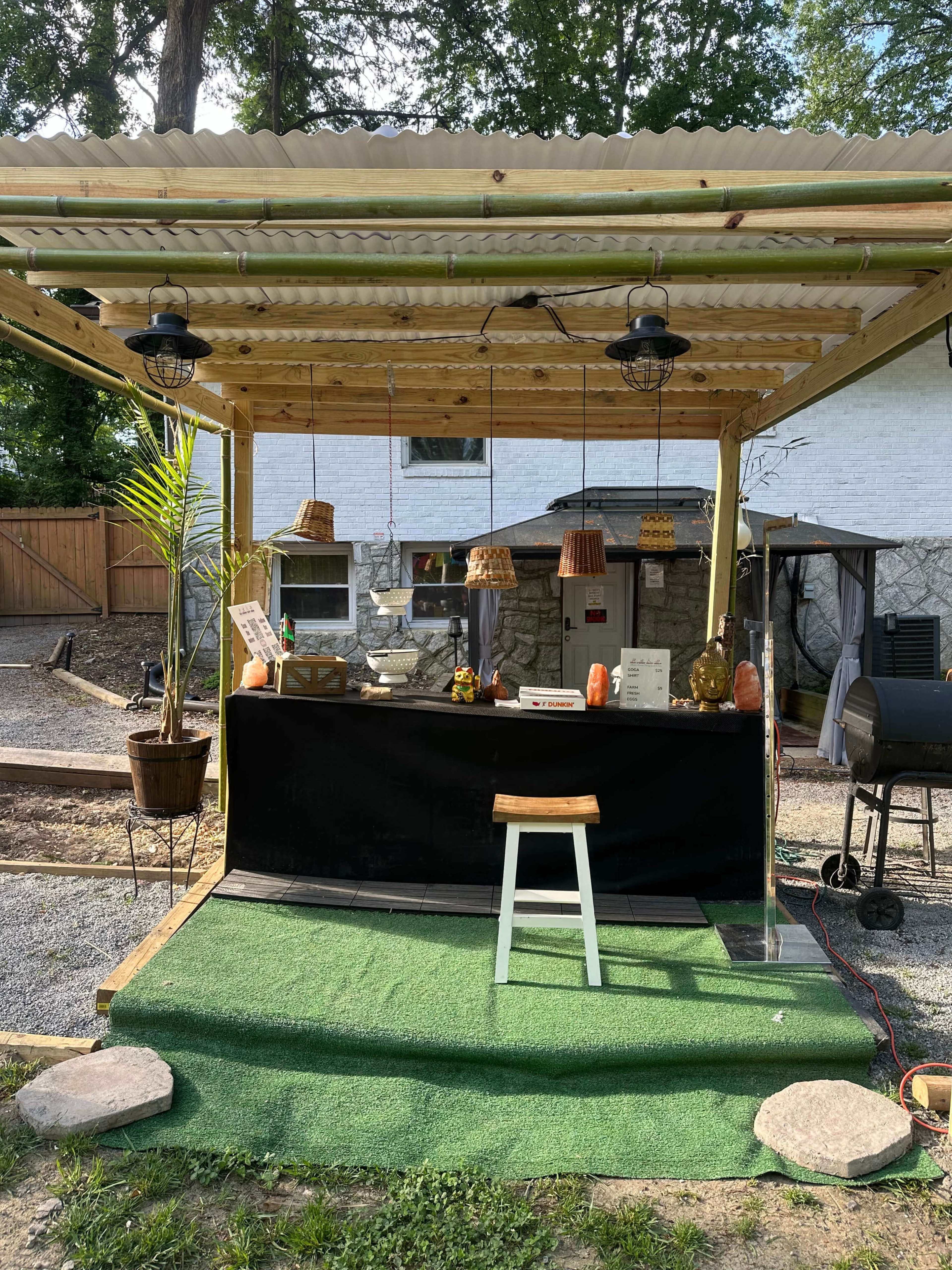 The image shows a small outdoor structure with a countertop, decorated with plants and a grill nearby, set against a backdrop of a house and gravel yard.