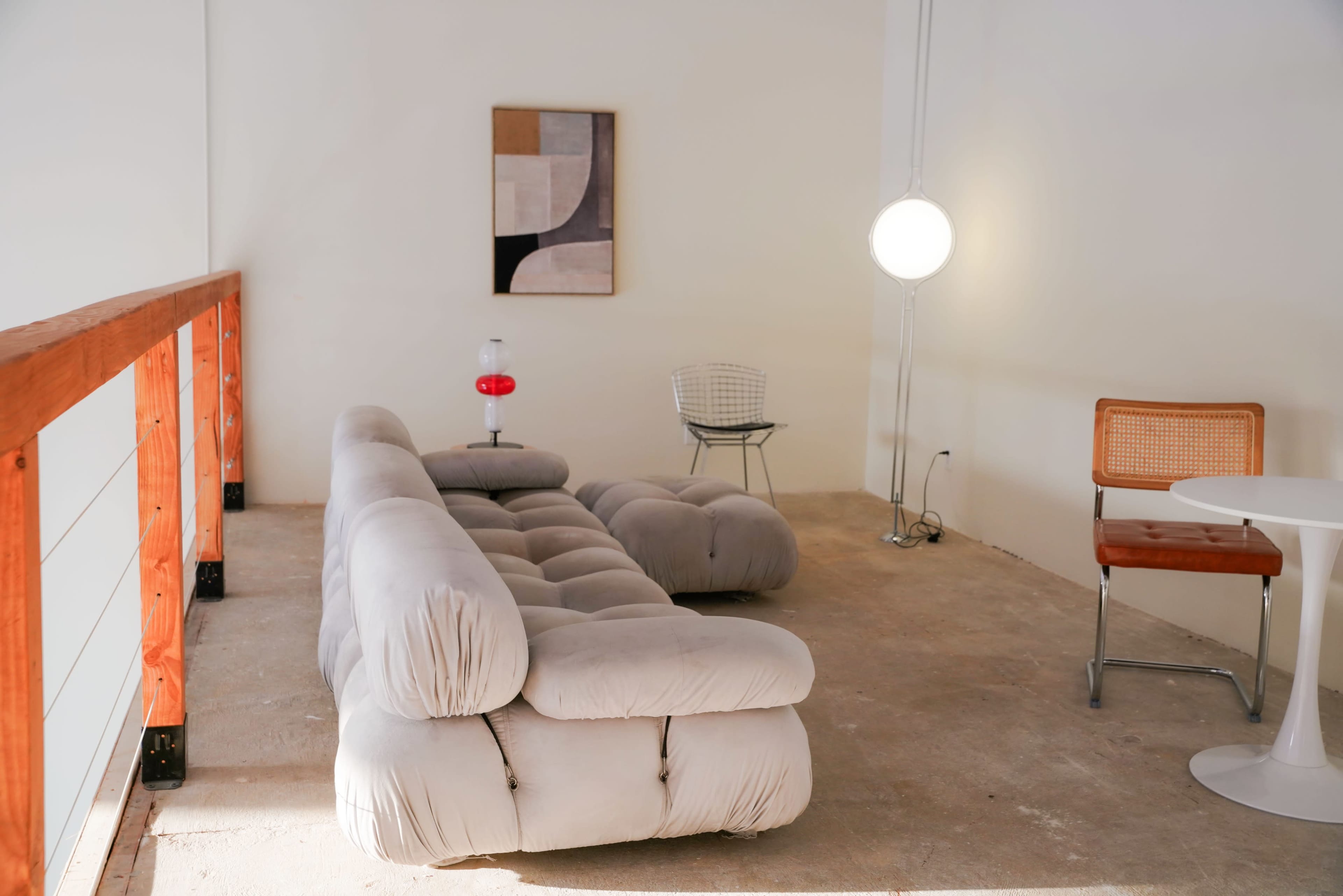 A modern interior features a gray sofa, a round table, a wire chair, and a floor lamp against a minimalist backdrop.