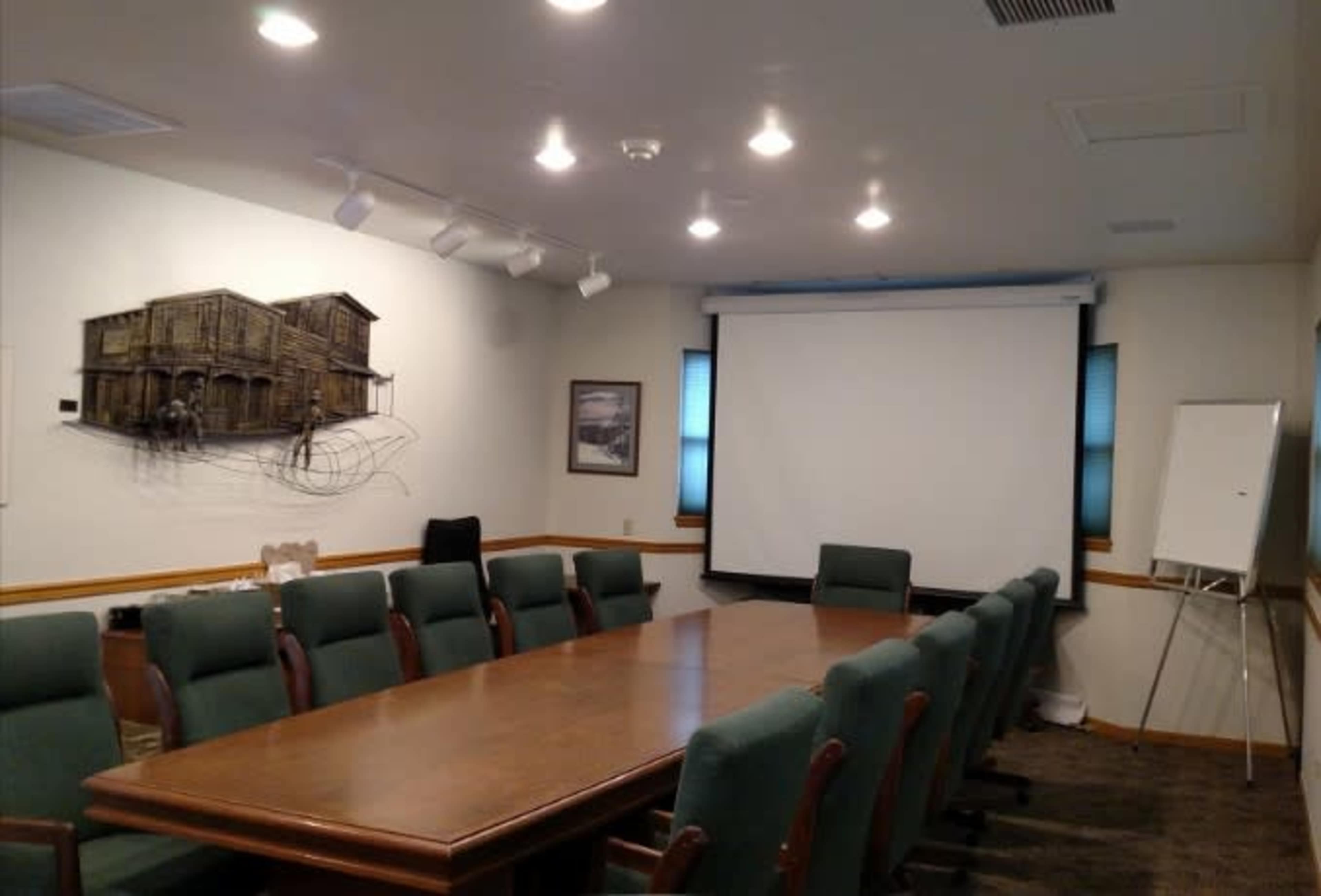 A conference room features a long wooden table surrounded by green chairs, with a projector screen and a whiteboard at one end.