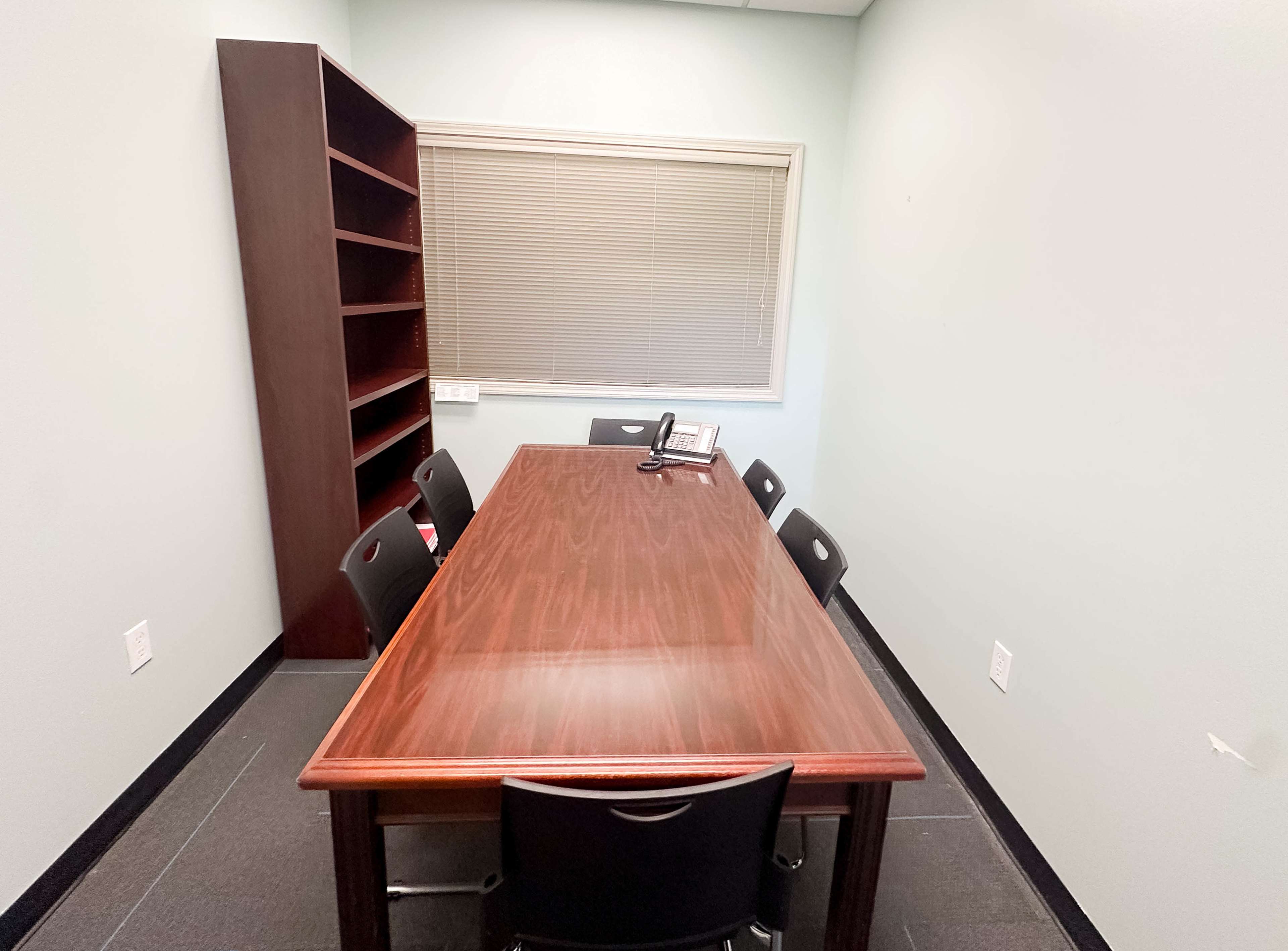 A rectangular conference table with six black chairs is centered in a small, empty room featuring a bookshelf and a telephone.