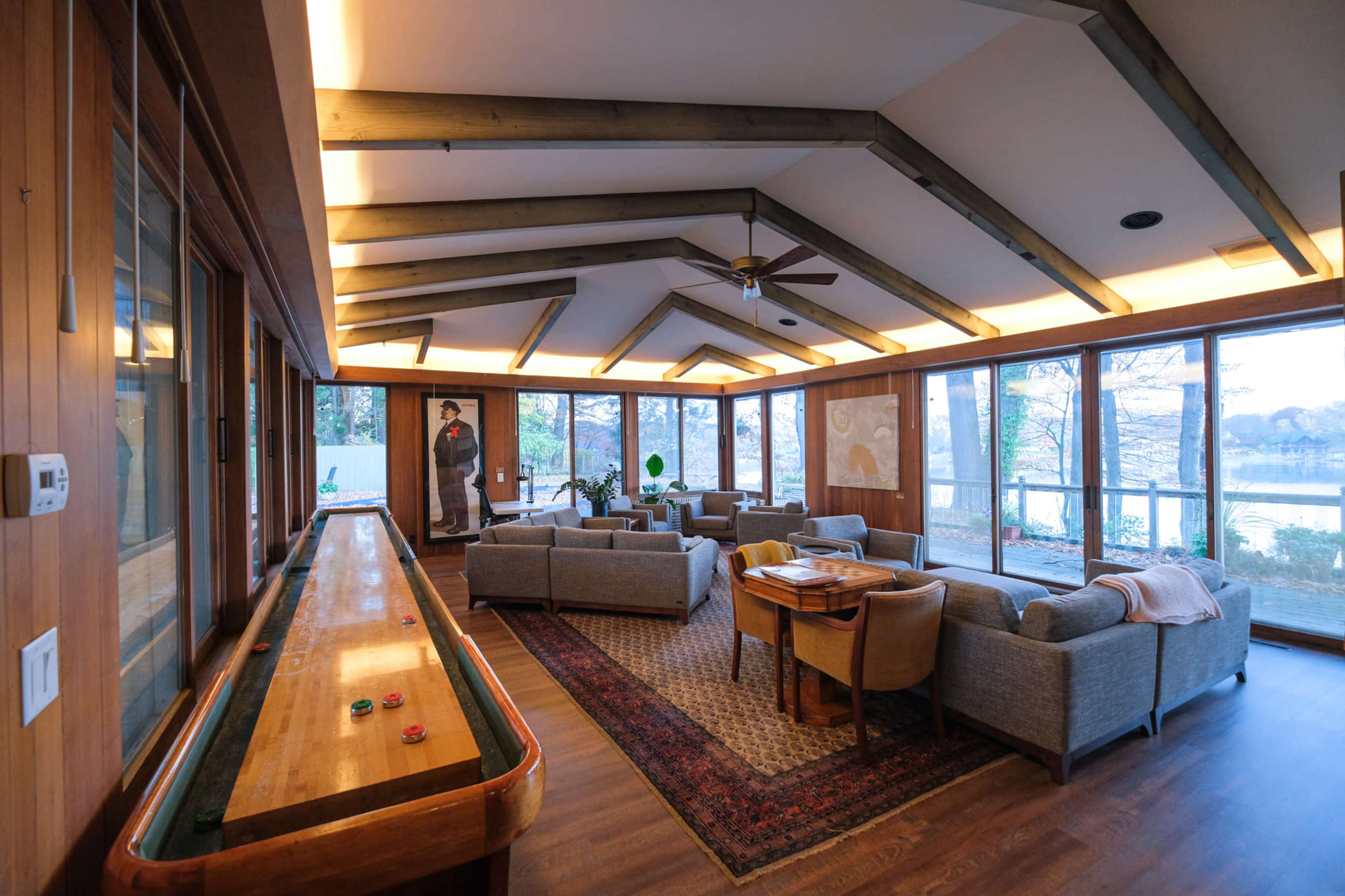 A spacious living area with large windows, exposed wooden beams, and a shuffleboard table along one side, overlooking a lake.