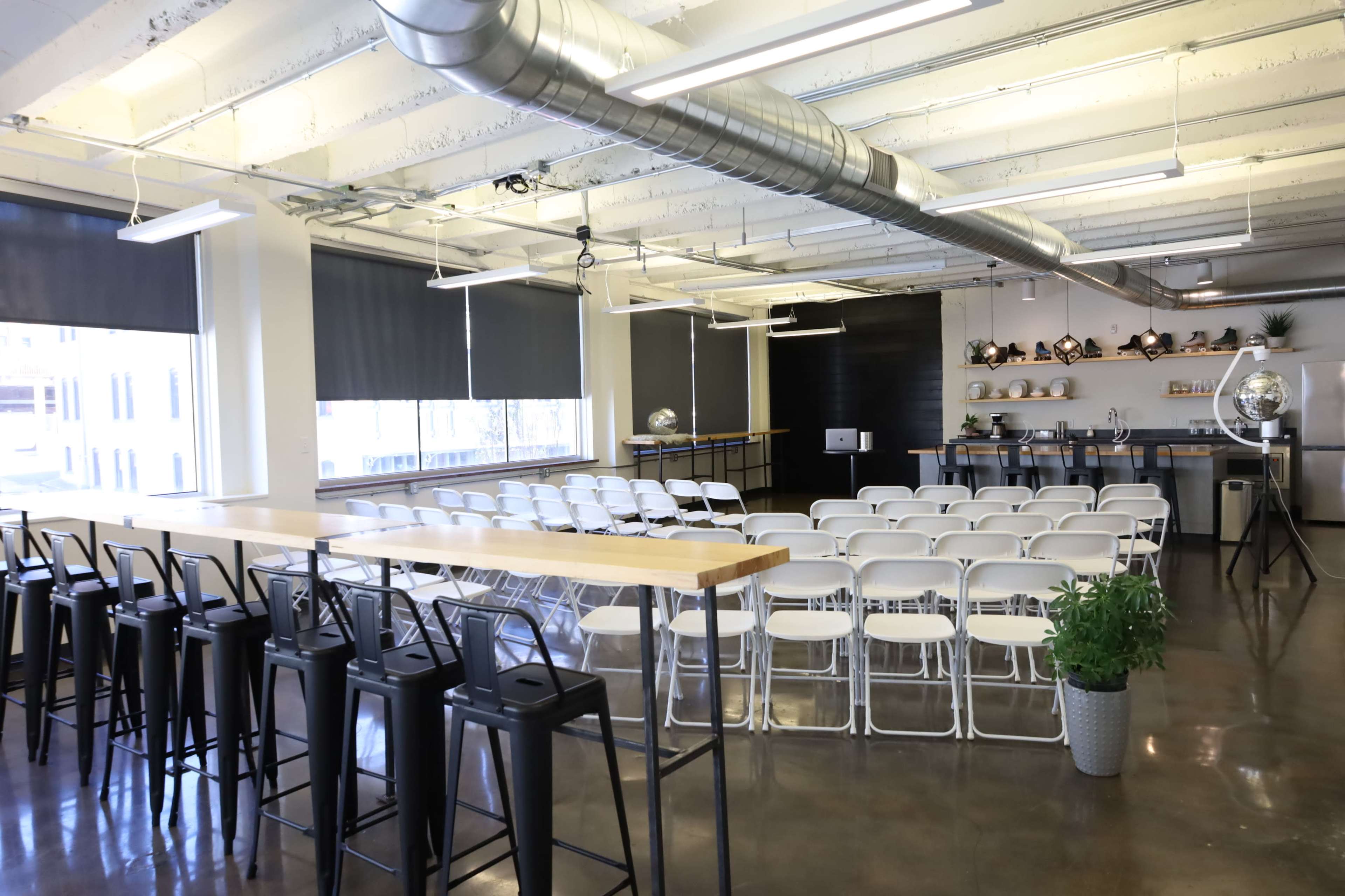 A modern event space with rows of white chairs facing a black wall, a high table along one side, and large windows allowing natural light to enter.