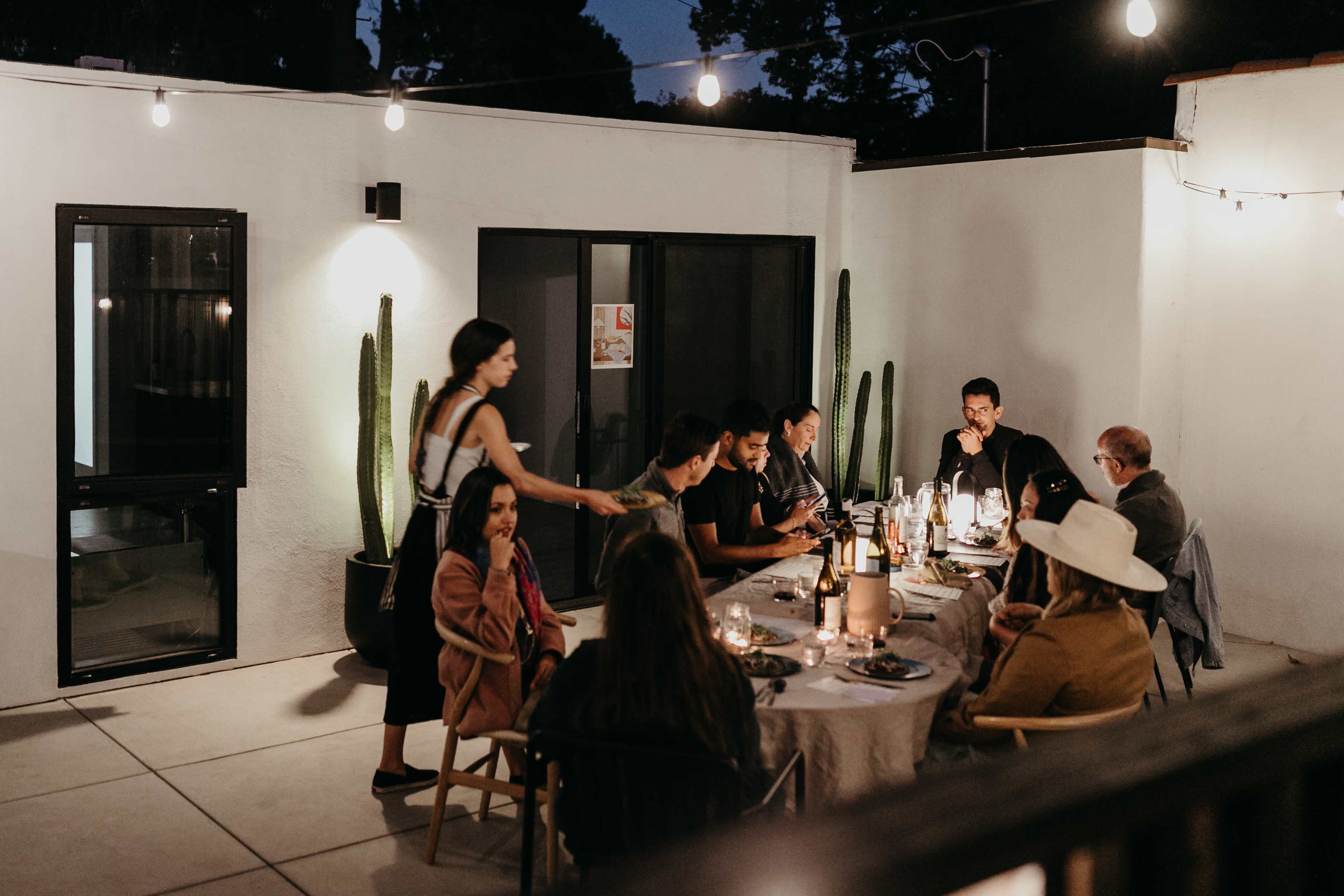 A group of people is gathered around a dining table on a patio at night, with string lights illuminating the scene.