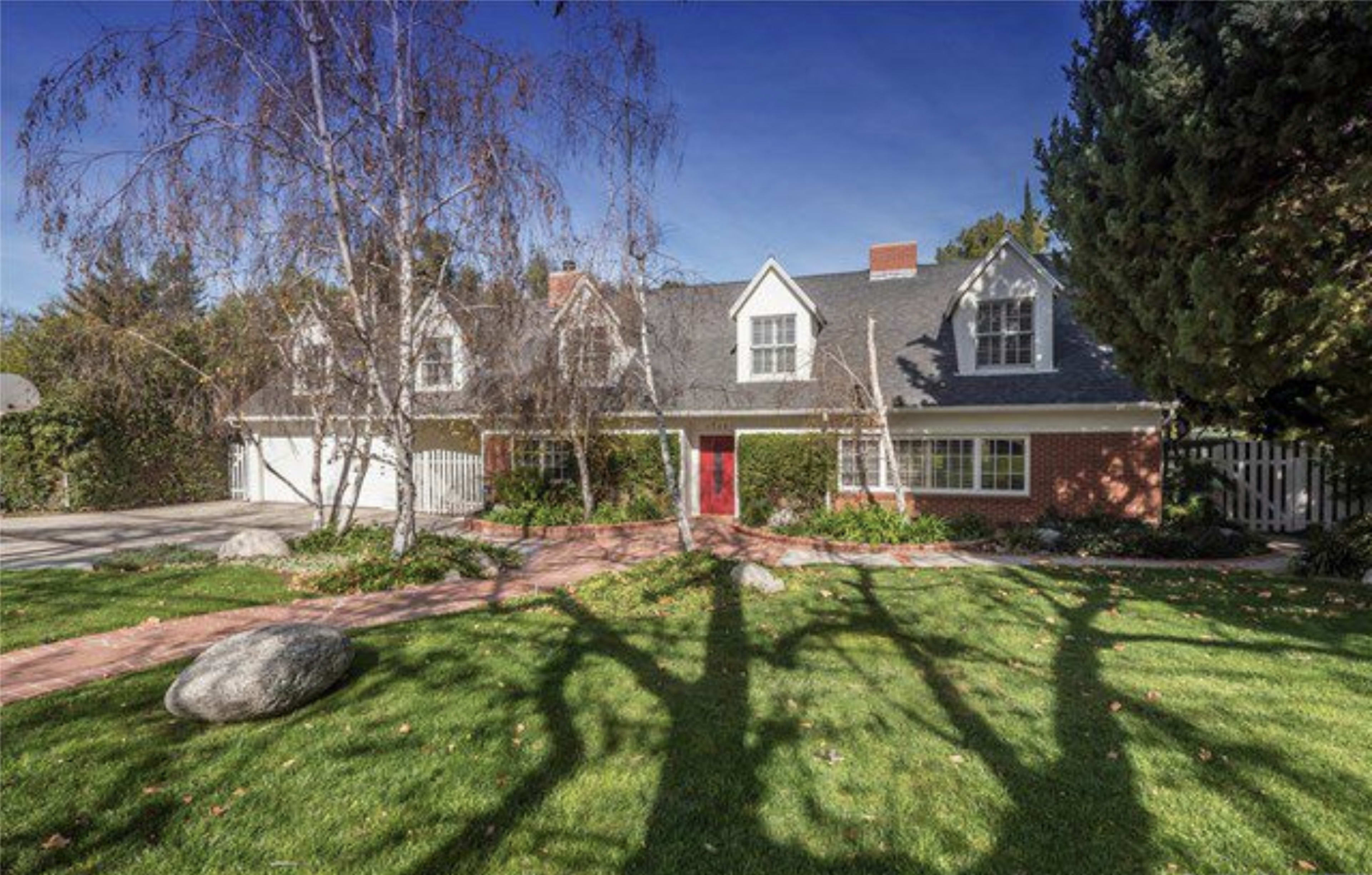 A two-story brick house with a gray roof and red door is surrounded by a landscaped yard featuring grass, shrubs, and a large rock.