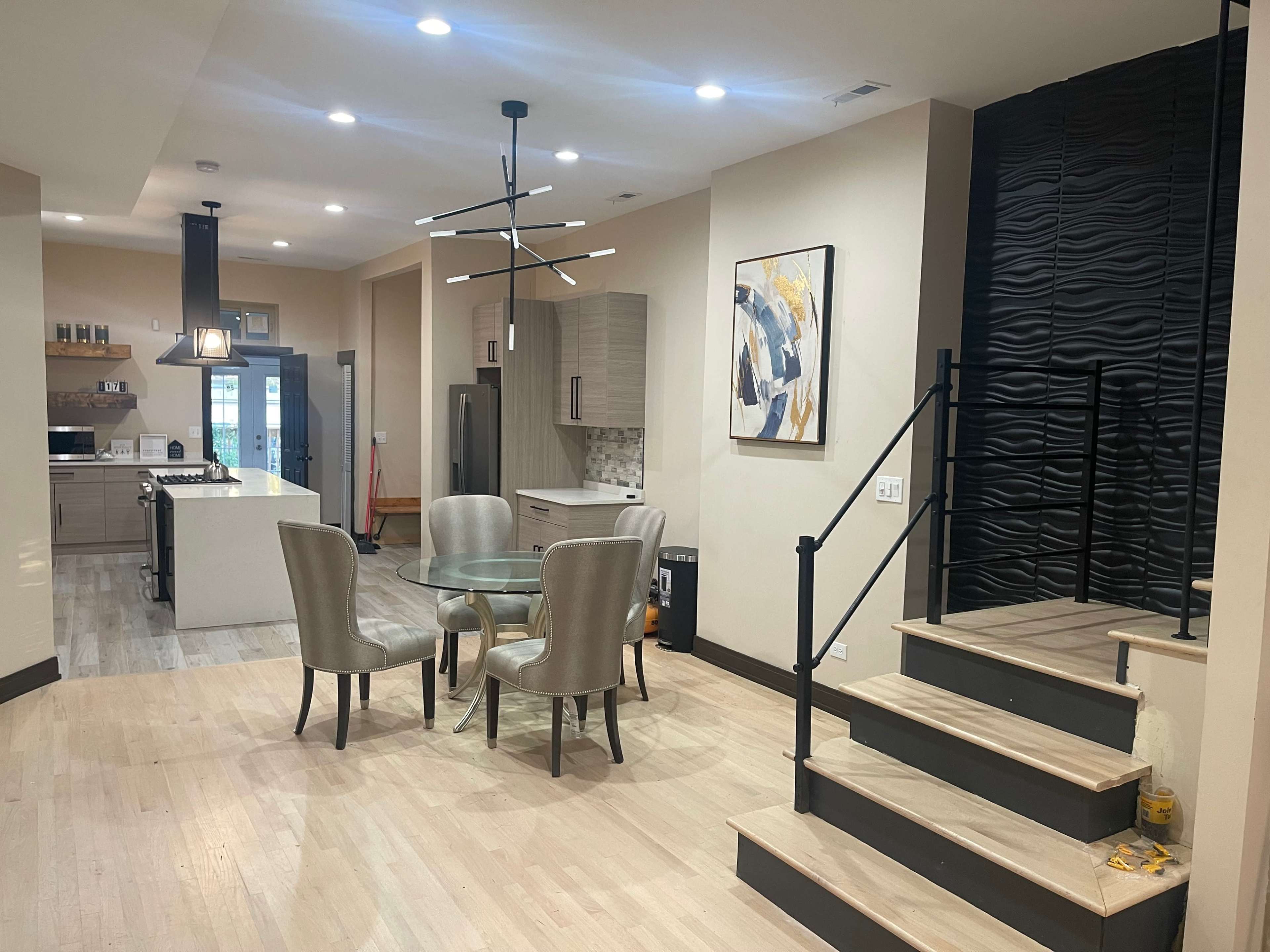 A modern interior features a dining area with a glass table and four chairs, leading to a kitchen with sleek cabinetry and an adjacent staircase.