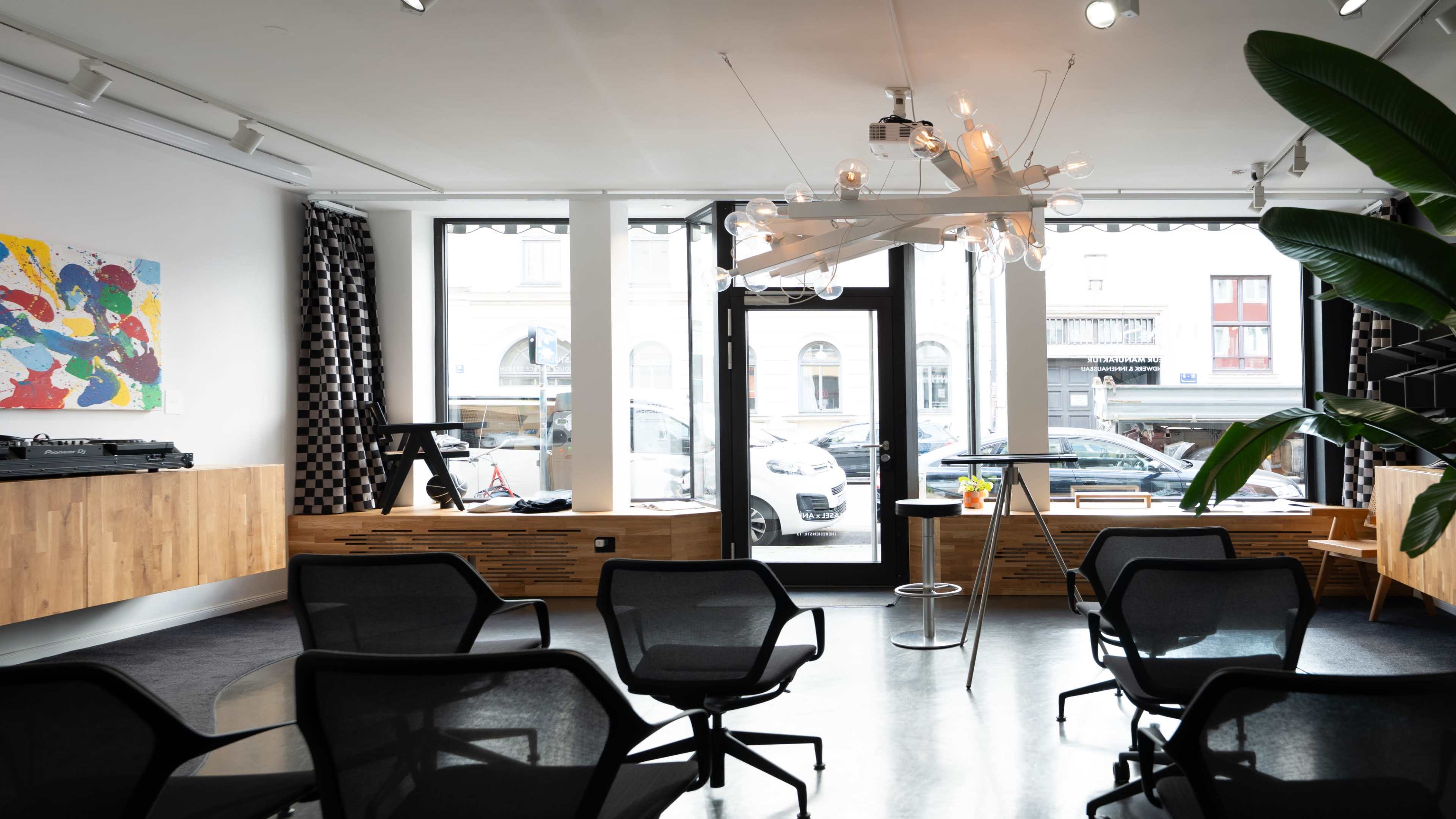 The image shows a modern, brightly lit interior of a space with wooden furniture, a large chandelier, and rows of black chairs arranged facing a window.
