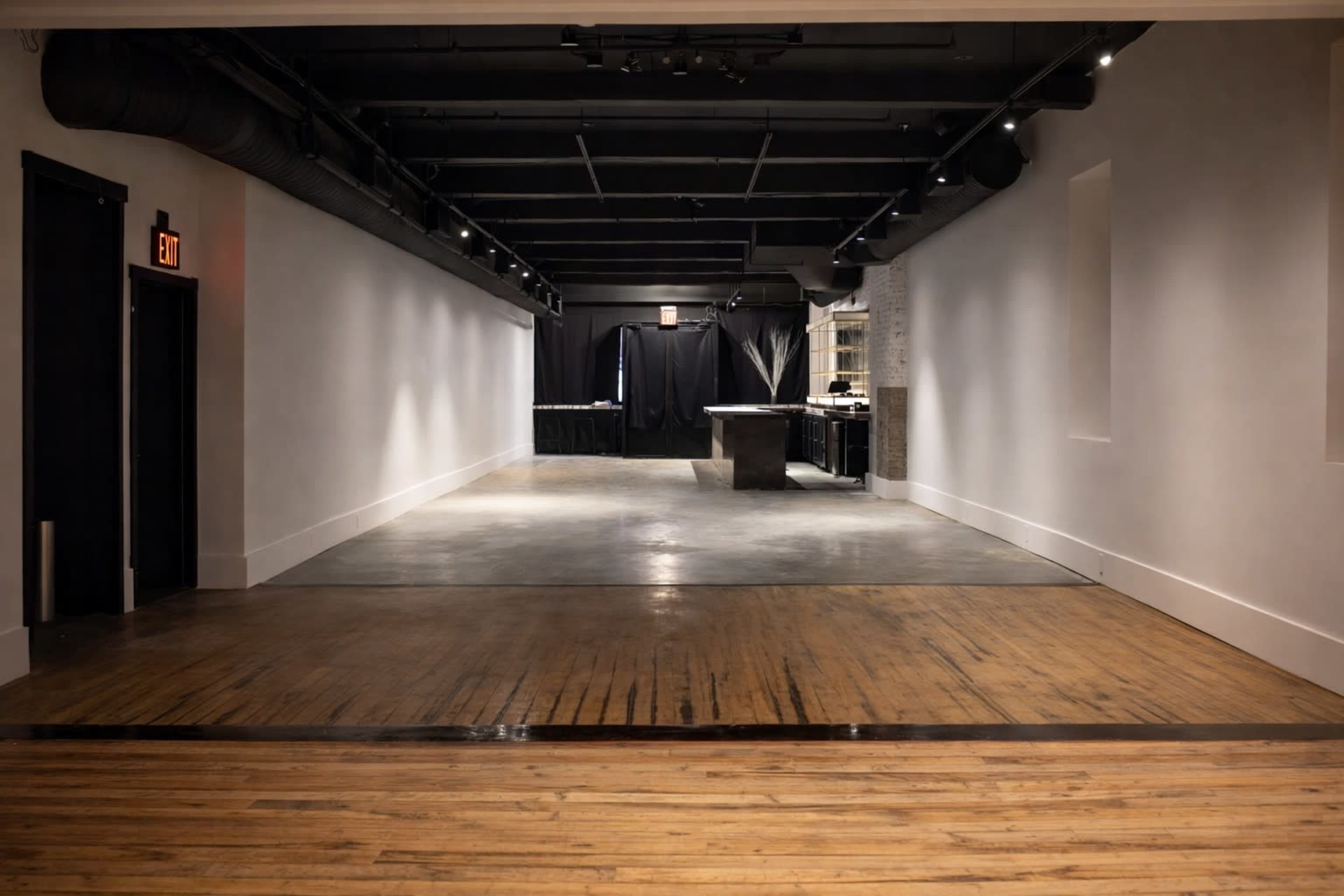 A spacious, well-lit interior features polished wooden floors and smooth gray concrete, leading to a darkened area with black drapery in the distance.