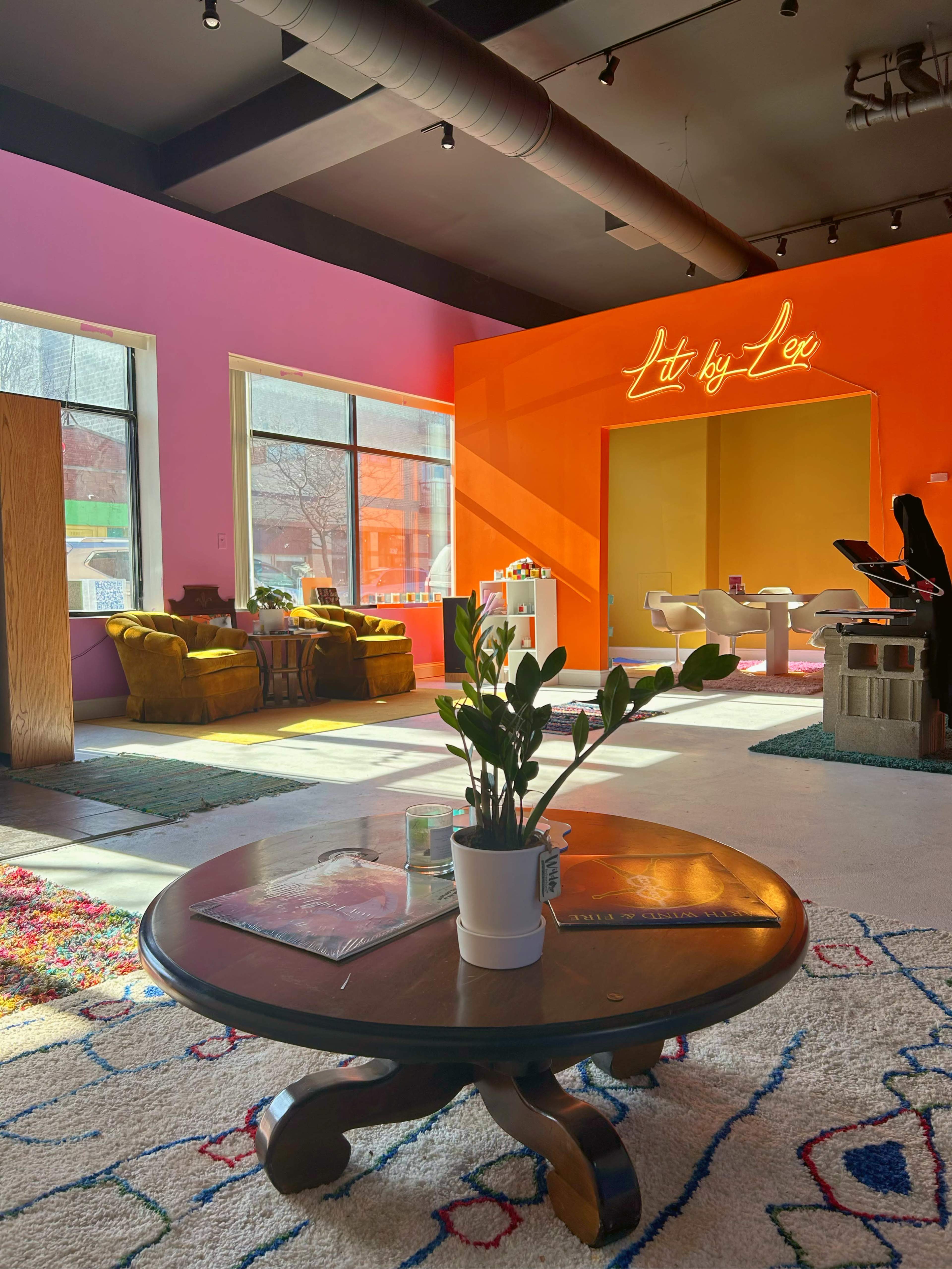 A brightly colored interior space with a pink wall, an orange wall labeled "Lit by Lex," a cozy seating area, and a small table with a plant in the center.