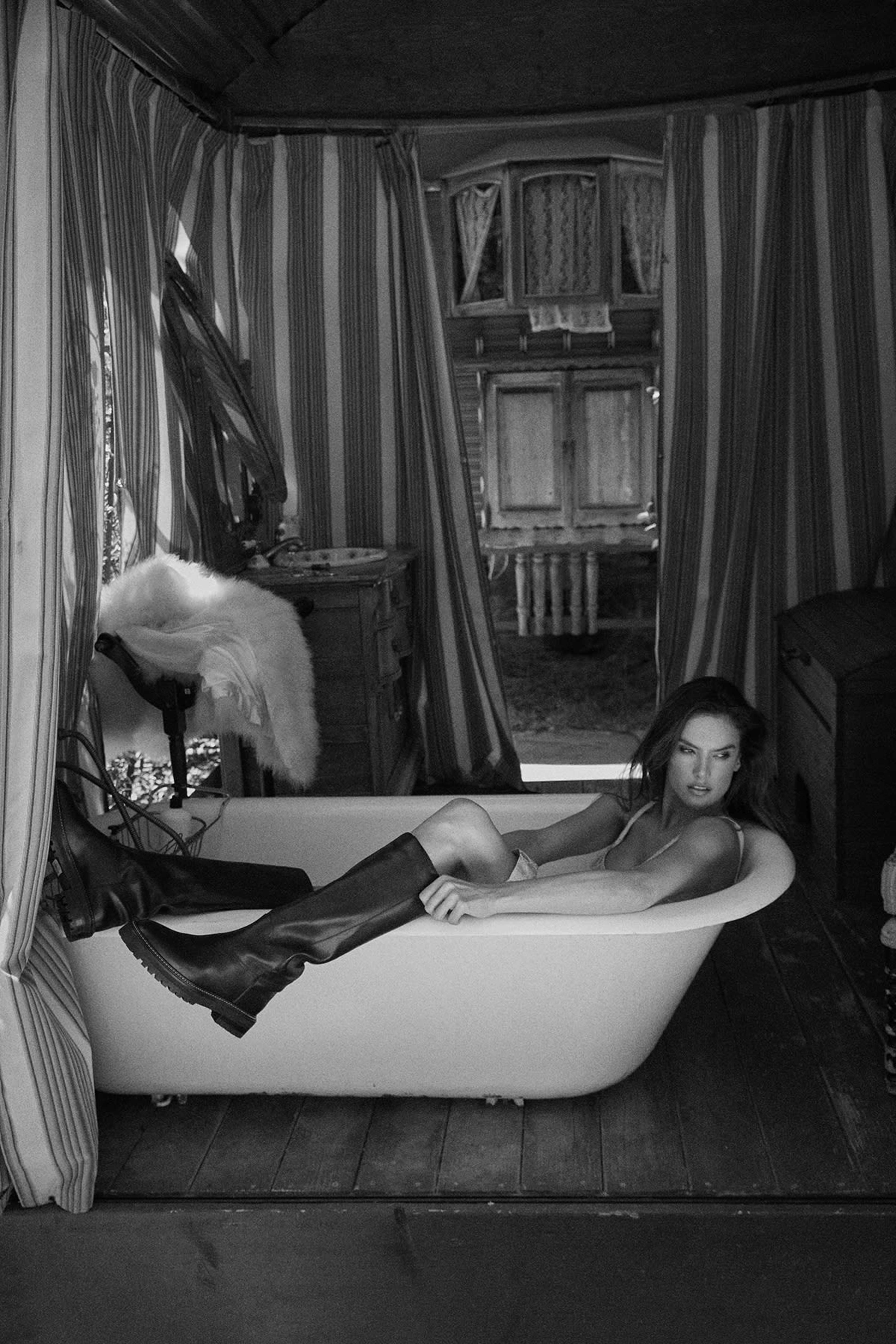 A person reclines in a vintage bathtub surrounded by striped curtains and rustic furniture.