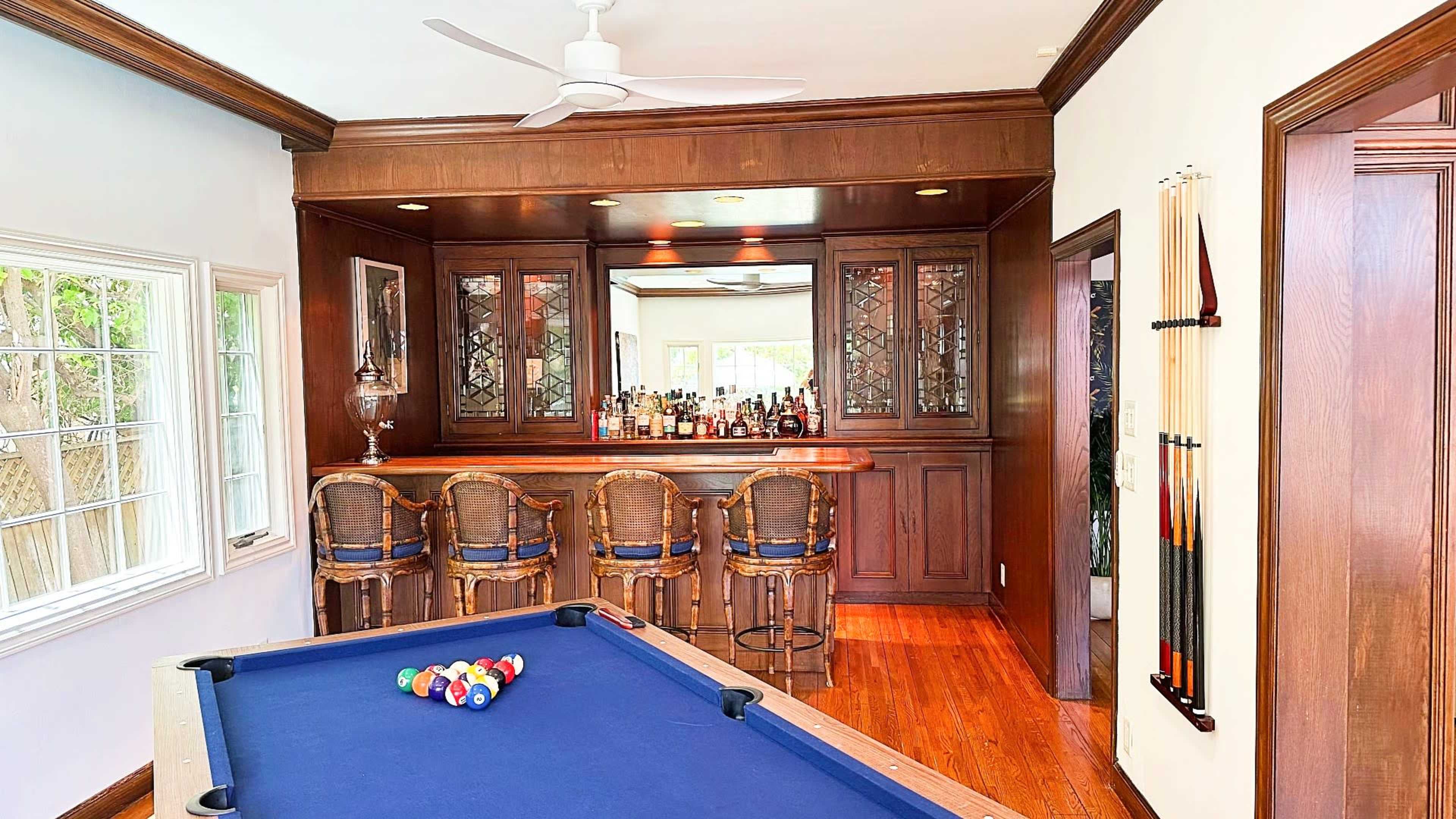 A room with a billiards table in the foreground and a bar area with seating and shelves filled with bottles in the background.