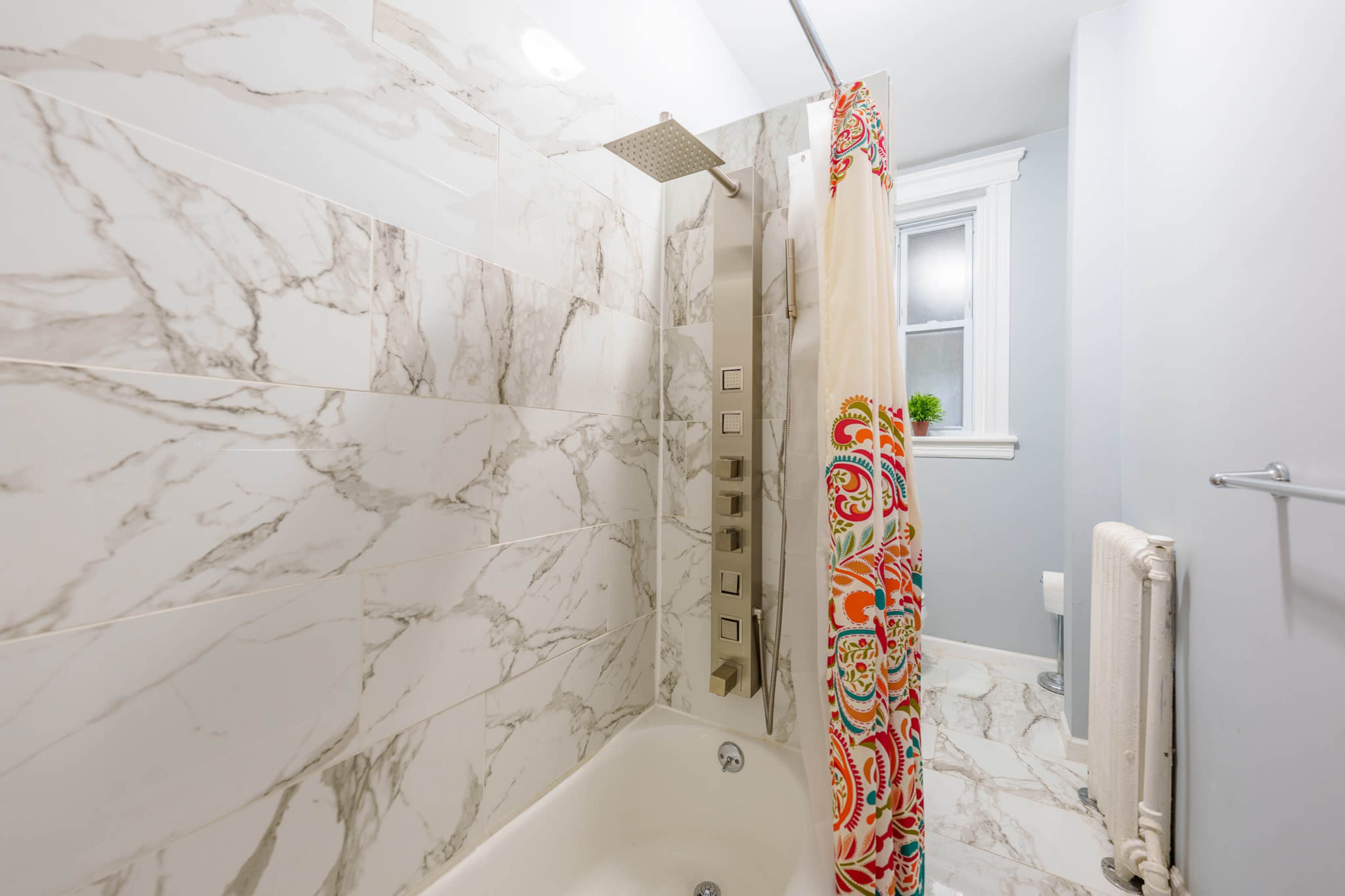 The image shows a bathroom with a marble-tiled shower, a colorful shower curtain, and a window allowing natural light.