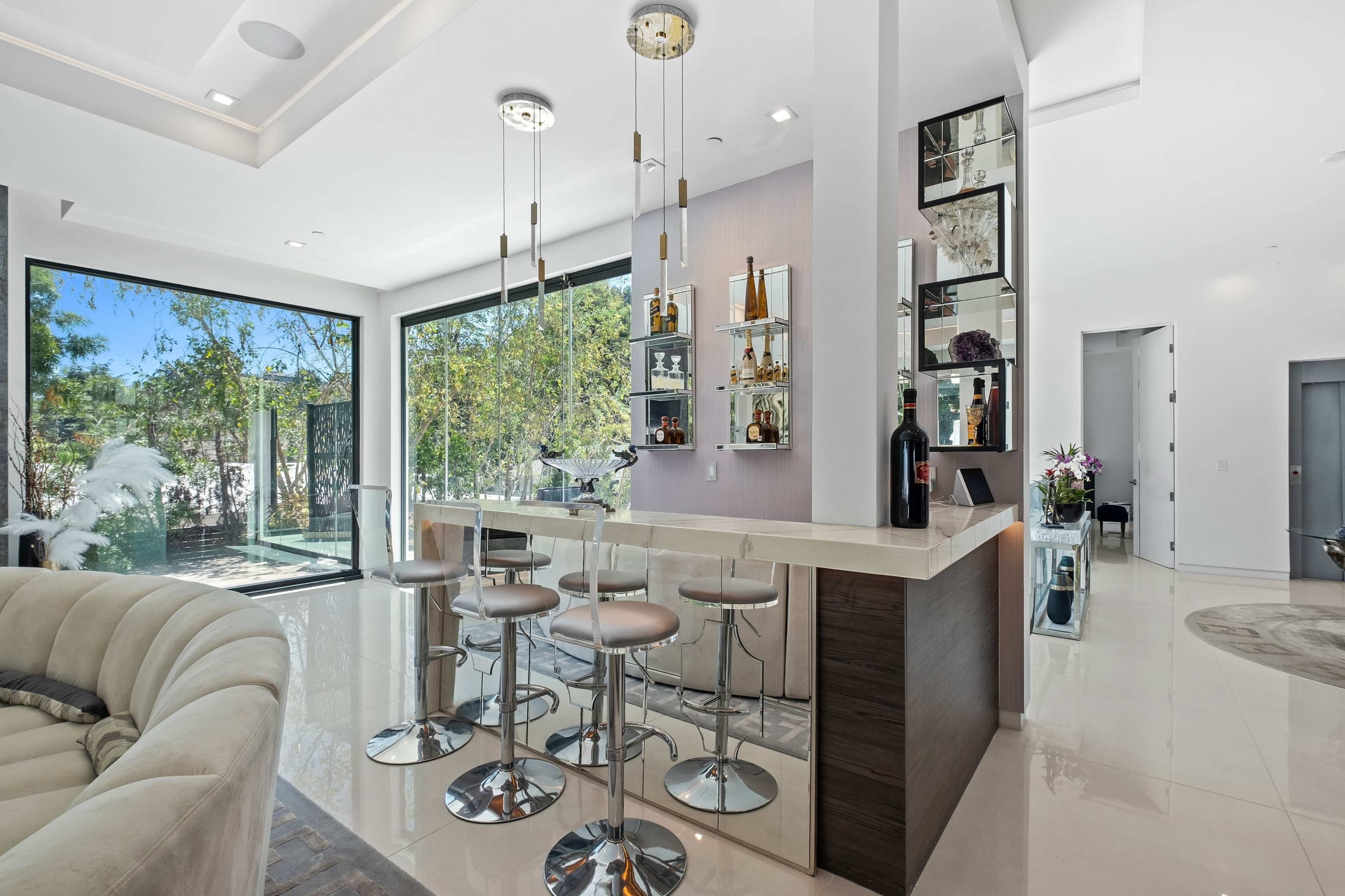 The image shows a modern kitchen with a bar area featuring stools, surrounded by large windows that provide views of greenery.