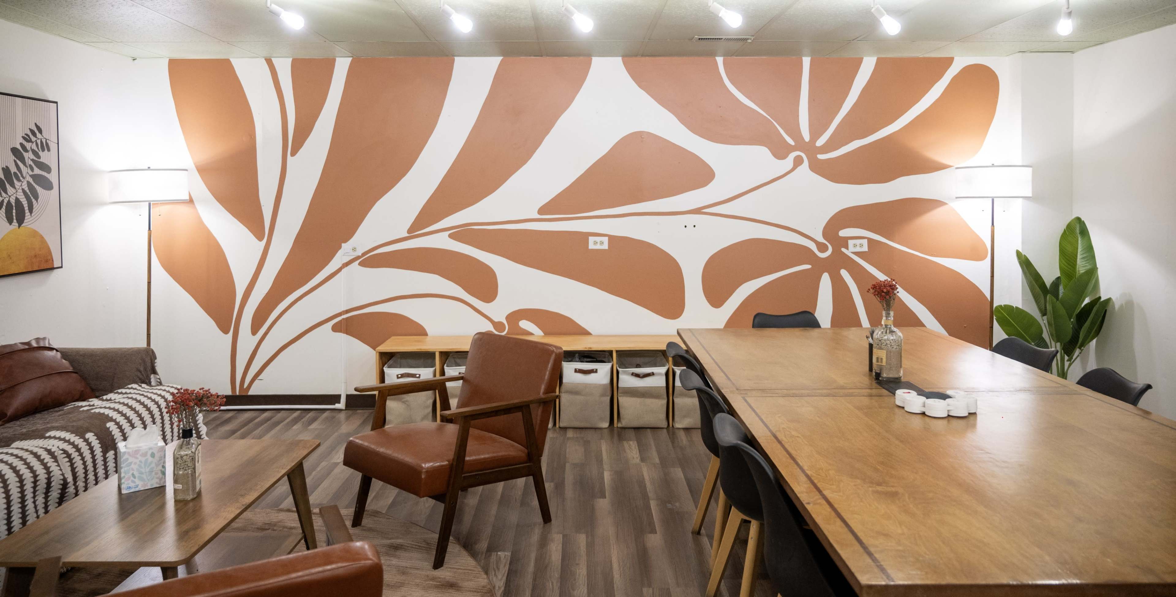 The image shows a cozy interior space with a large wooden table, several chairs, a sofa, and a mural of orange abstract shapes on the wall.