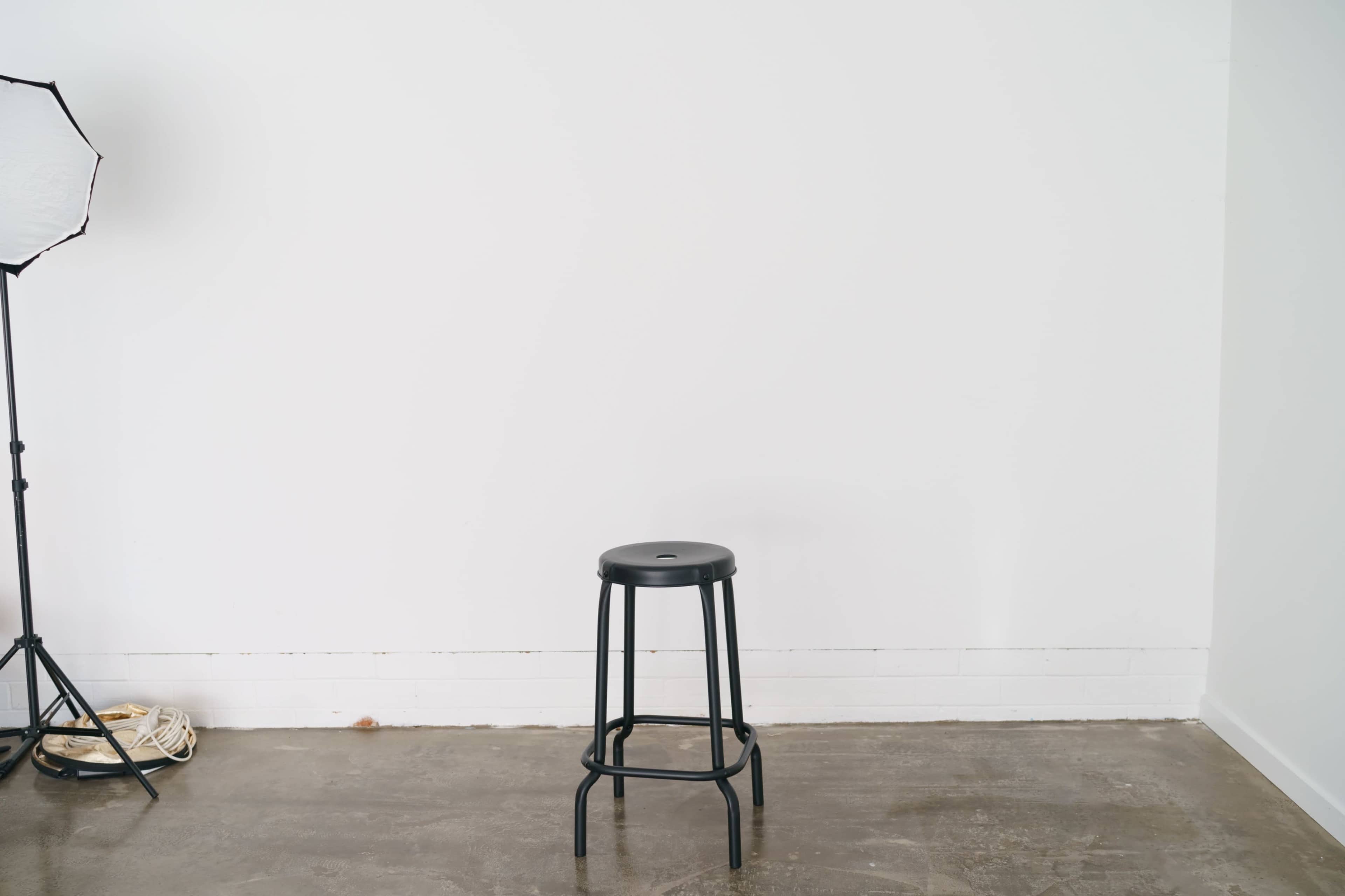 A black stool stands in the center of a minimalist room with a plain white wall and a photography light stand to the left.