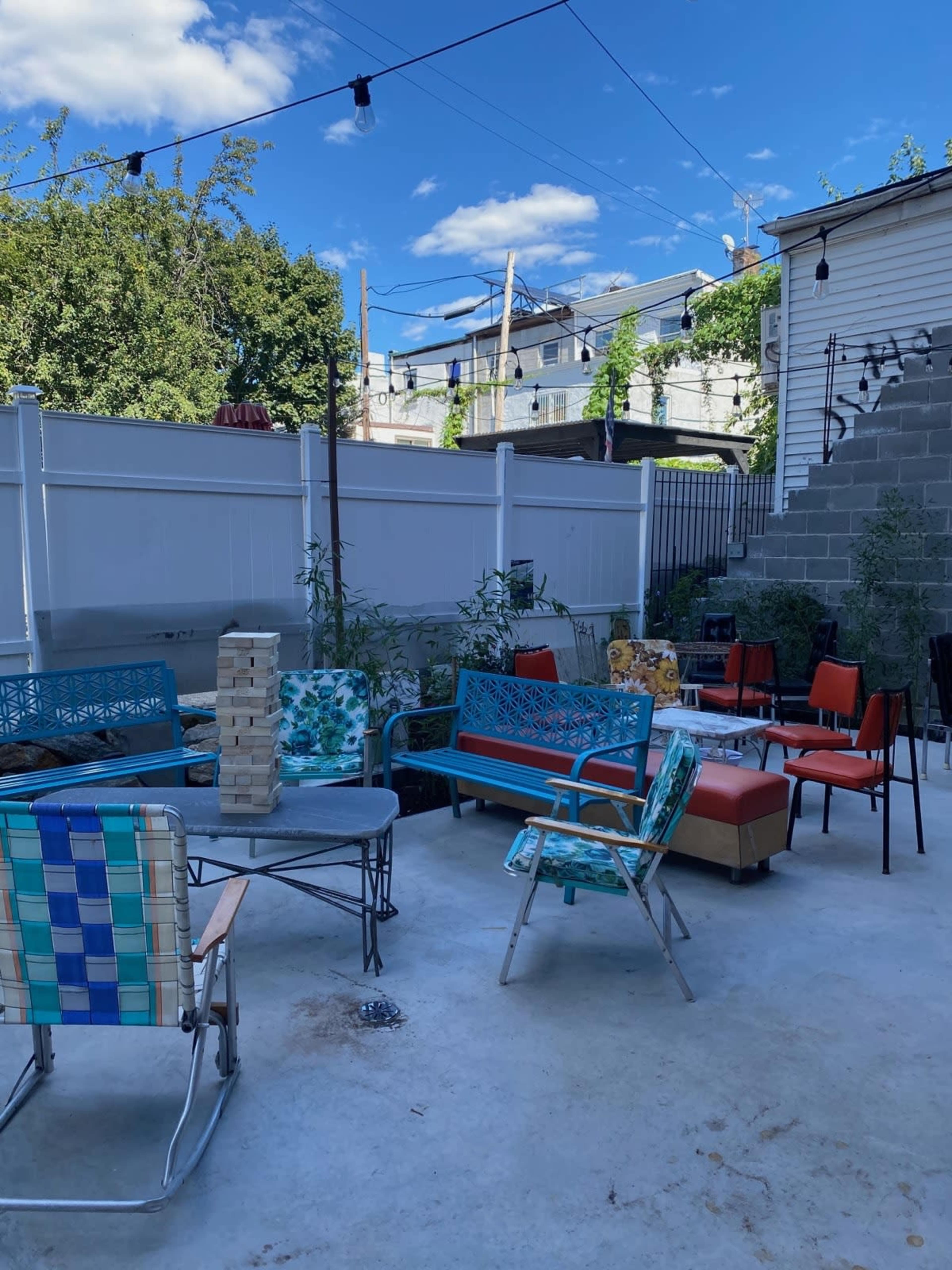 The image shows a backyard seating area featuring various colorful chairs and tables, with string lights overhead and a white fence in the background.
