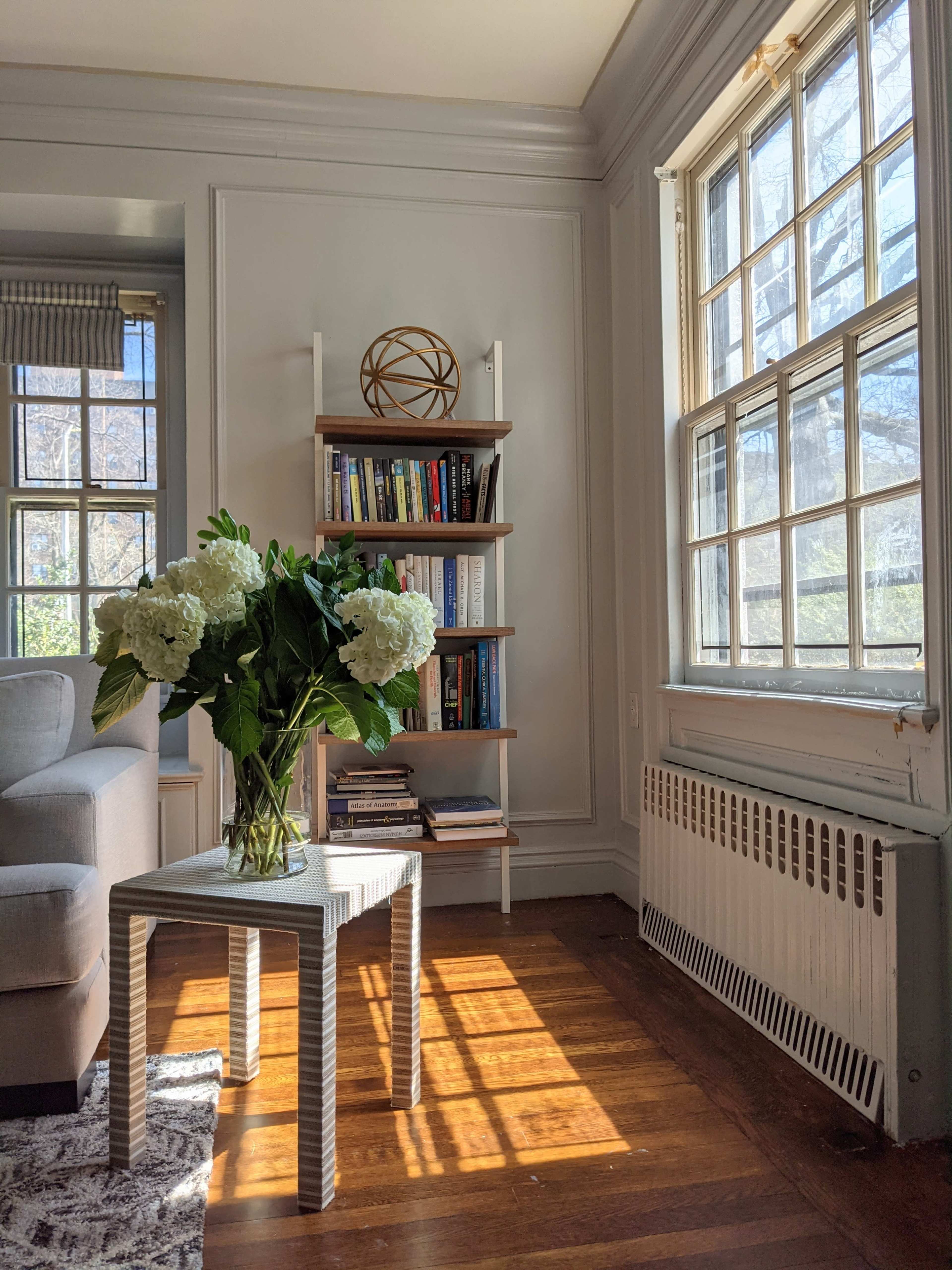 A small table with a vase of white flowers sits beside a bookshelf filled with books in a sunlit corner of a living room.