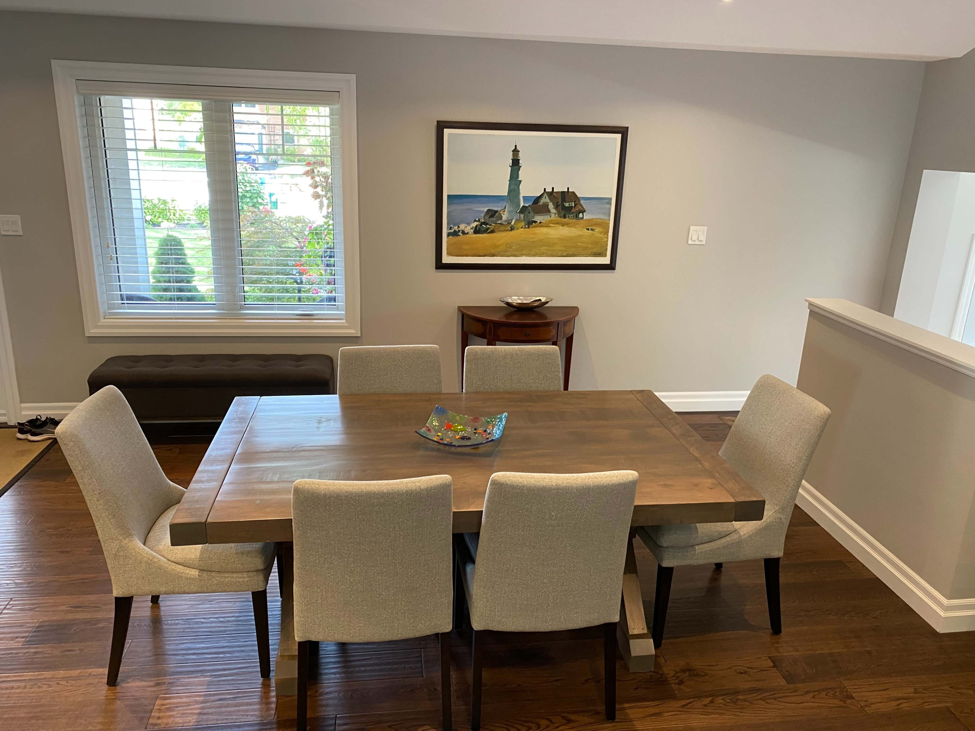 A wooden dining table surrounded by four upholstered chairs is positioned in a well-lit room, with a painting of a lighthouse on the wall and a console table in the background.