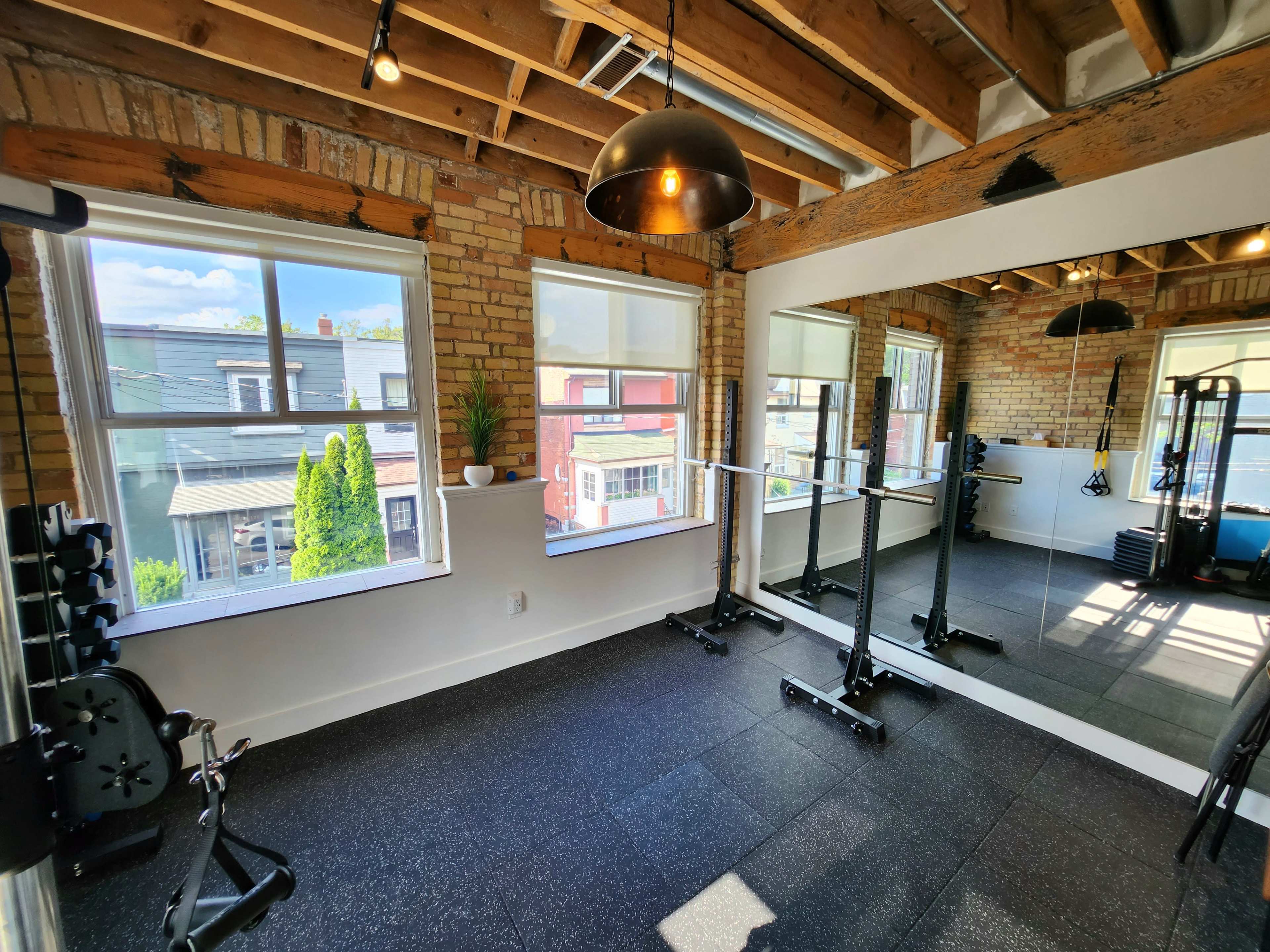 A bright gym with large windows, exposed brick walls, and a mirror, featuring weightlifting equipment and a rubber floor.