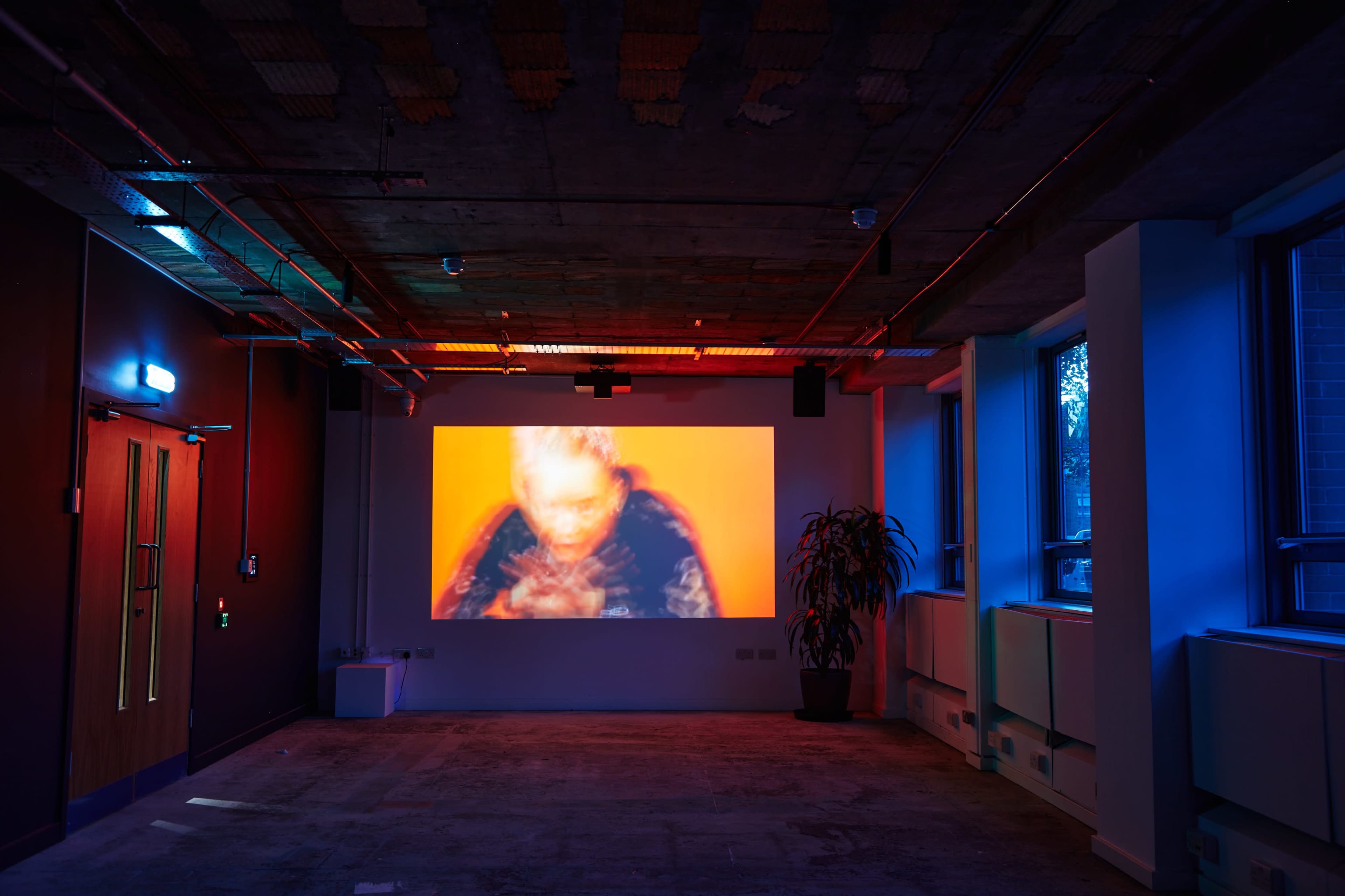 A dimly lit room features a large projection on the wall, displaying a blurred image of a figure, with colorful lighting illuminating the space.