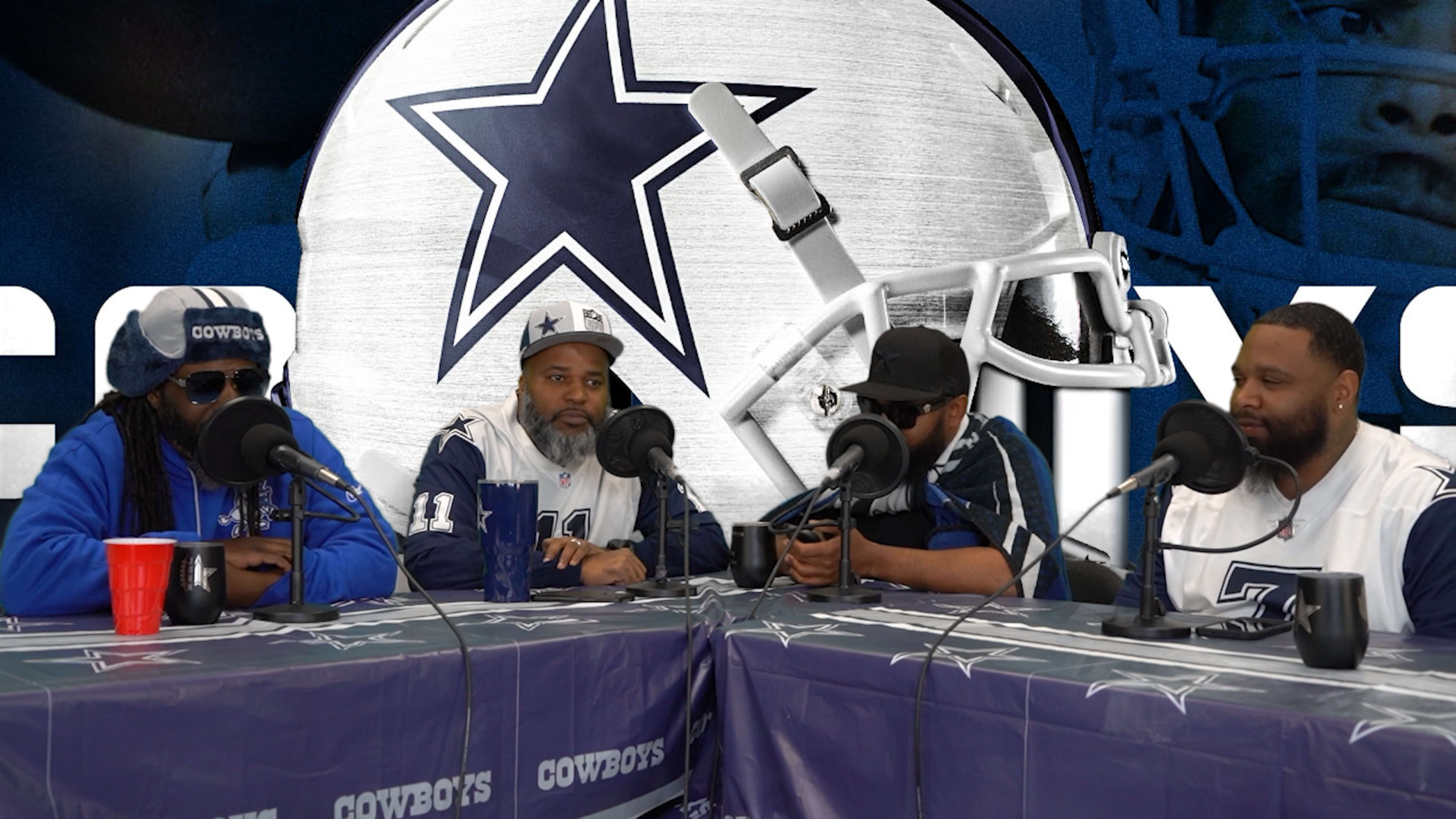 A group of four individuals wearing Dallas Cowboys gear are seated at a table with microphones, discussing in front of a large Cowboys logo backdrop.