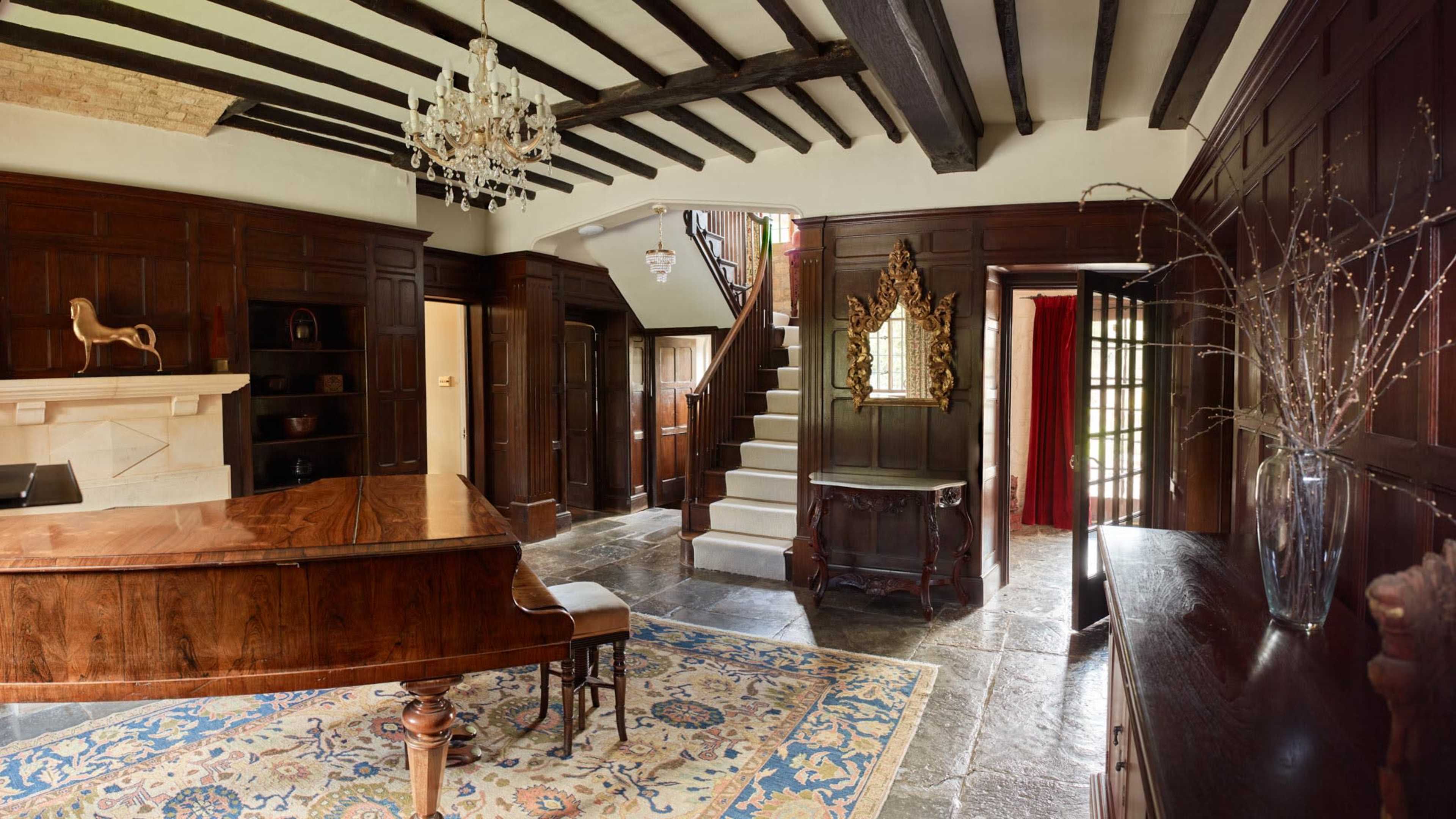 An elegant interior of a room featuring dark wooden paneling, a grand piano, a staircase, and a chandelier overhead.