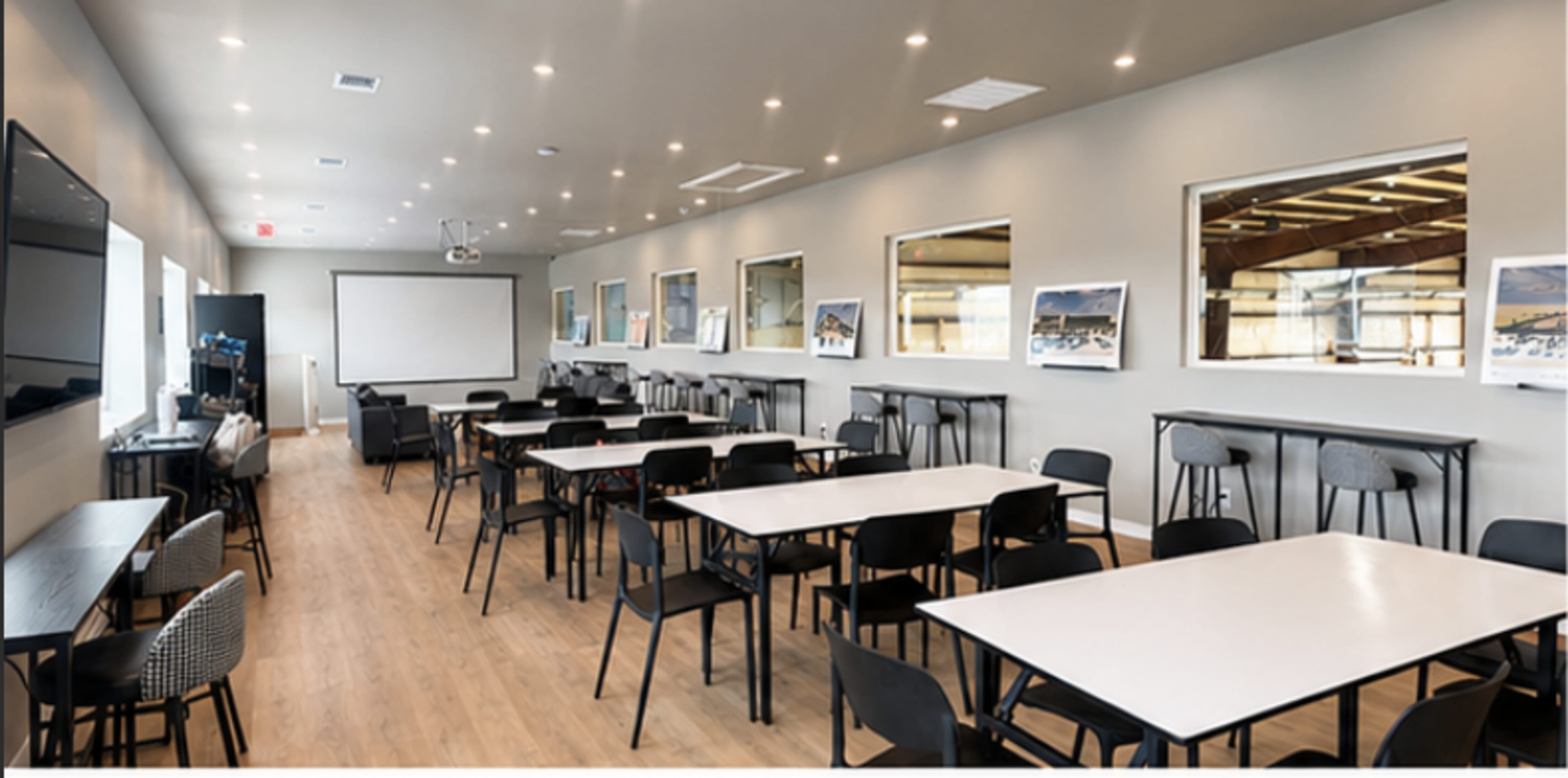 The image shows a spacious meeting room with several tables and chairs arranged for seating, featuring a large screen at one end and windows along the walls.