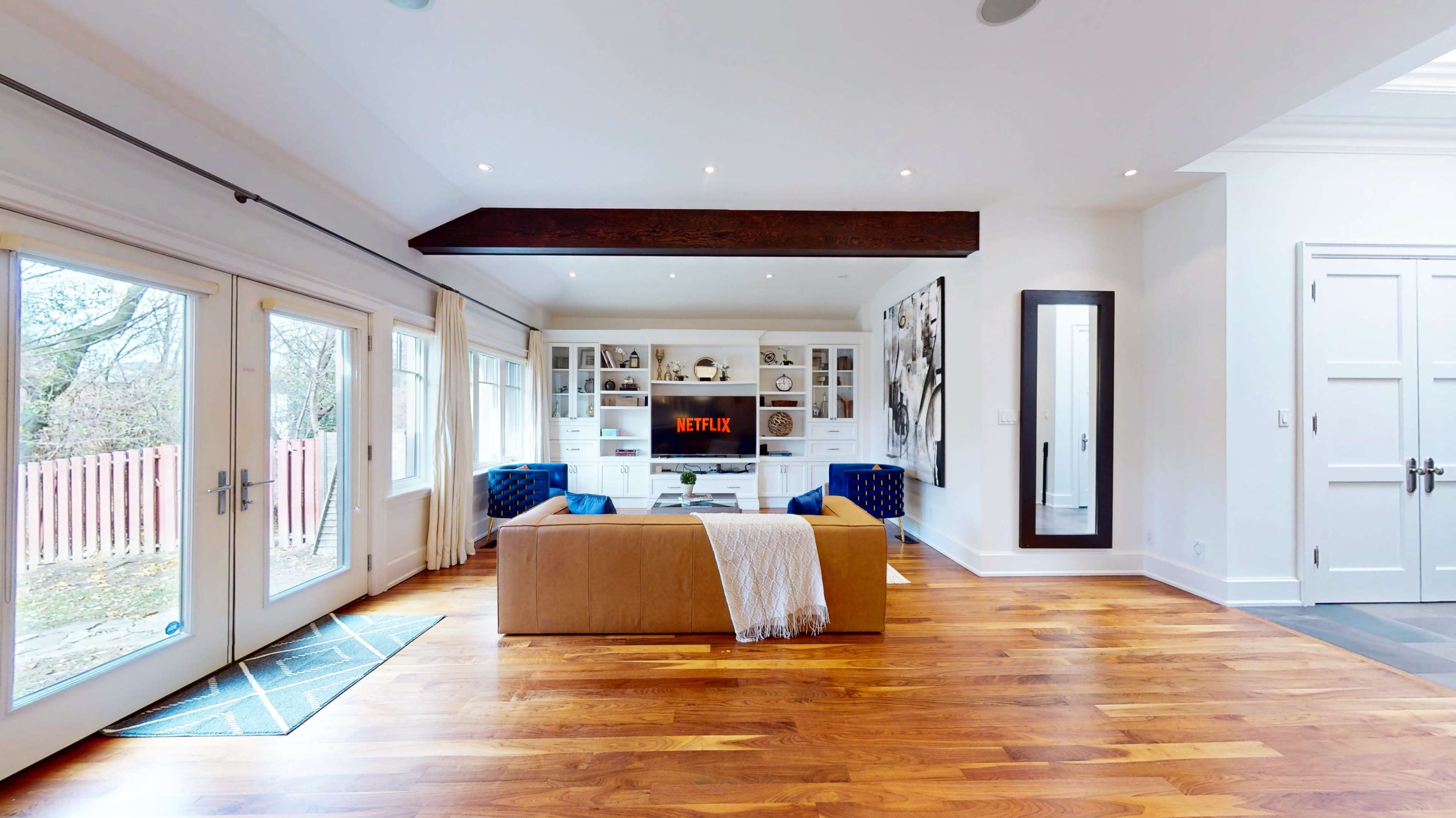 The living room features a modern layout with a beige sofa, a coffee table, shelves behind it, and a large television displaying the Netflix logo, all set against wooden flooring and natural light from large windows.