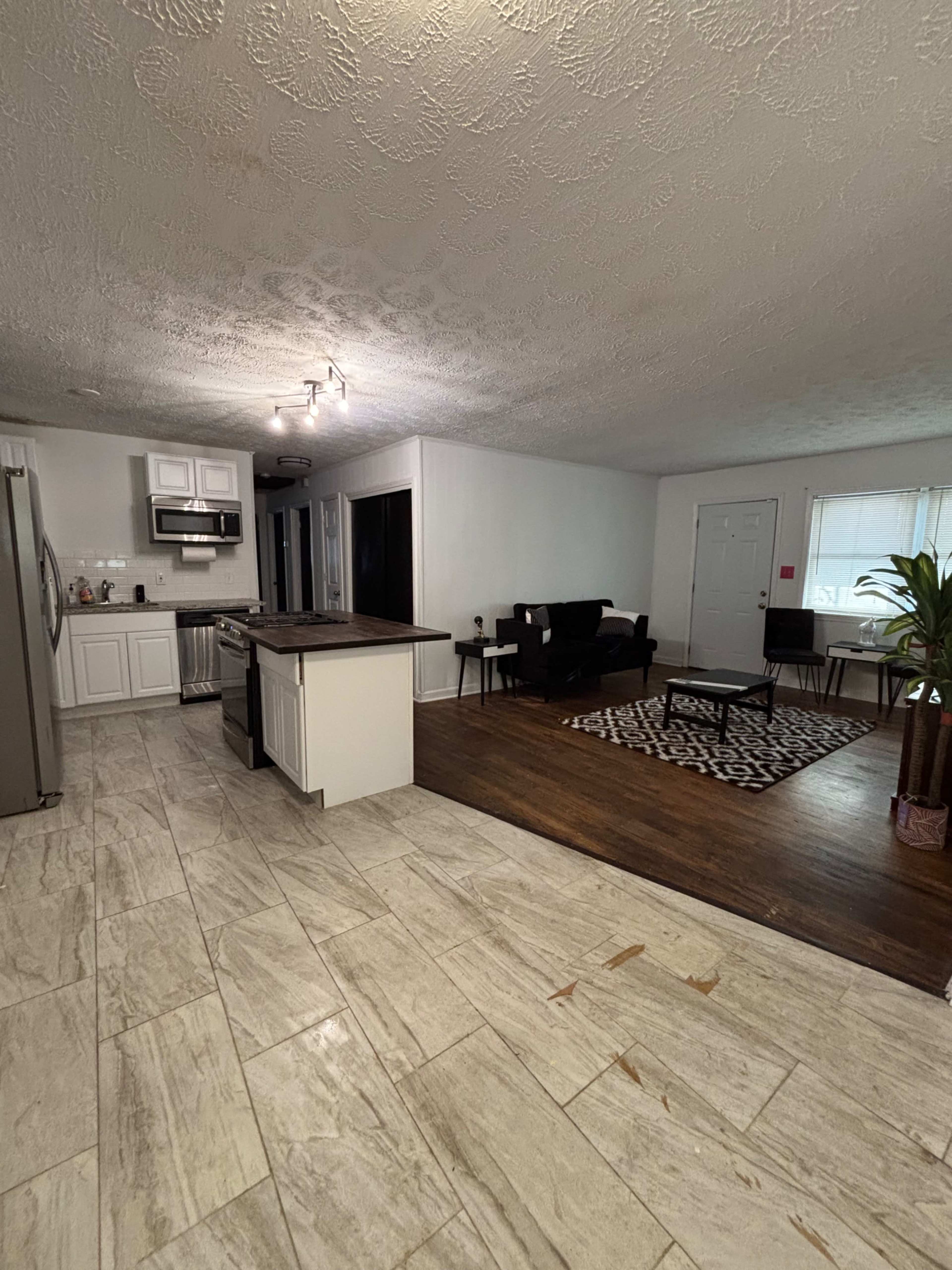 The image shows a spacious living area with a kitchen on one side, featuring tiled floors, a wooden accent area, and various seating arrangements.