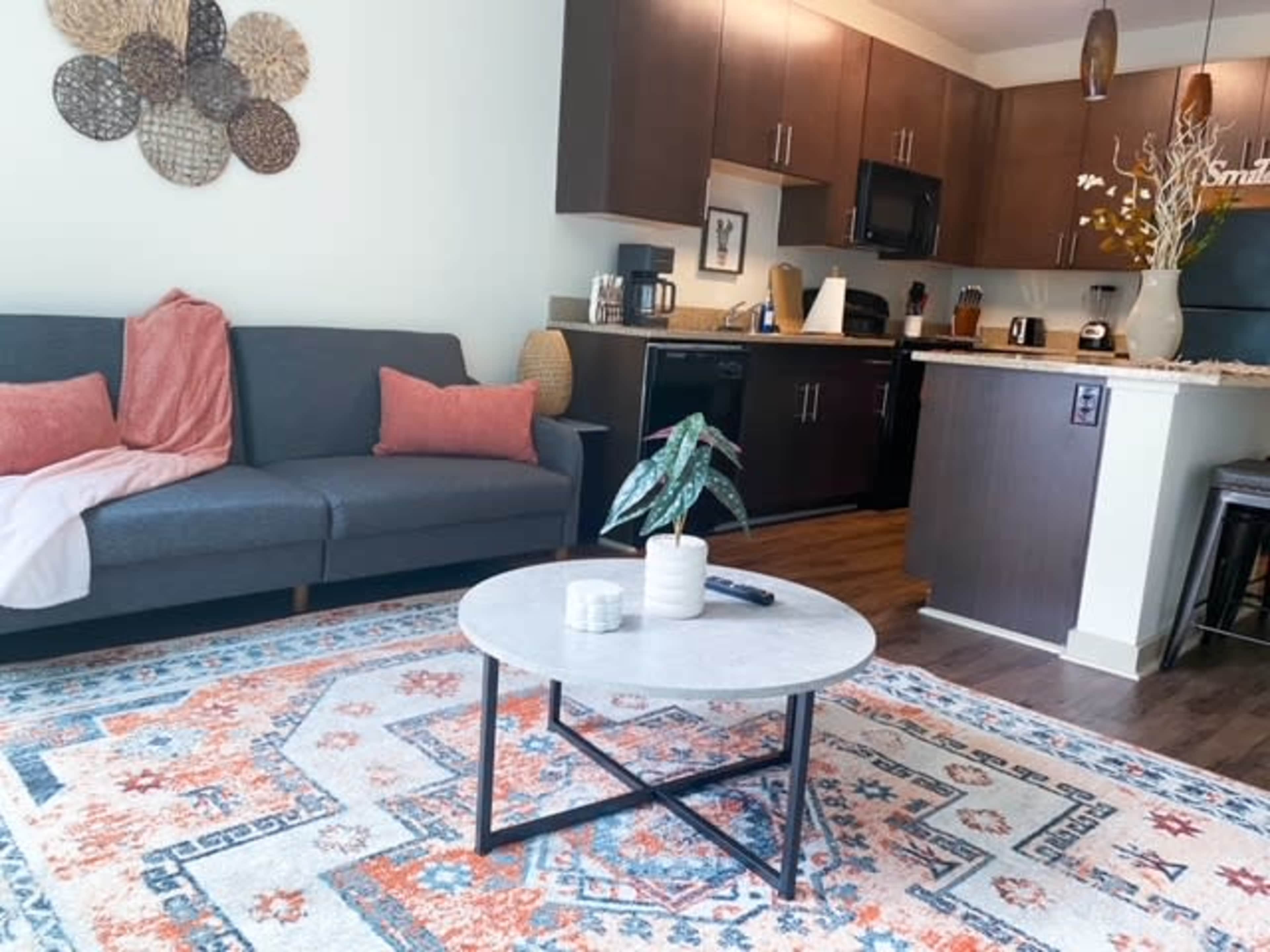 A cozy living room features a gray sofa with pink cushions, a marble-top coffee table, and a colorful area rug, adjacent to a modern kitchen with dark cabinetry.