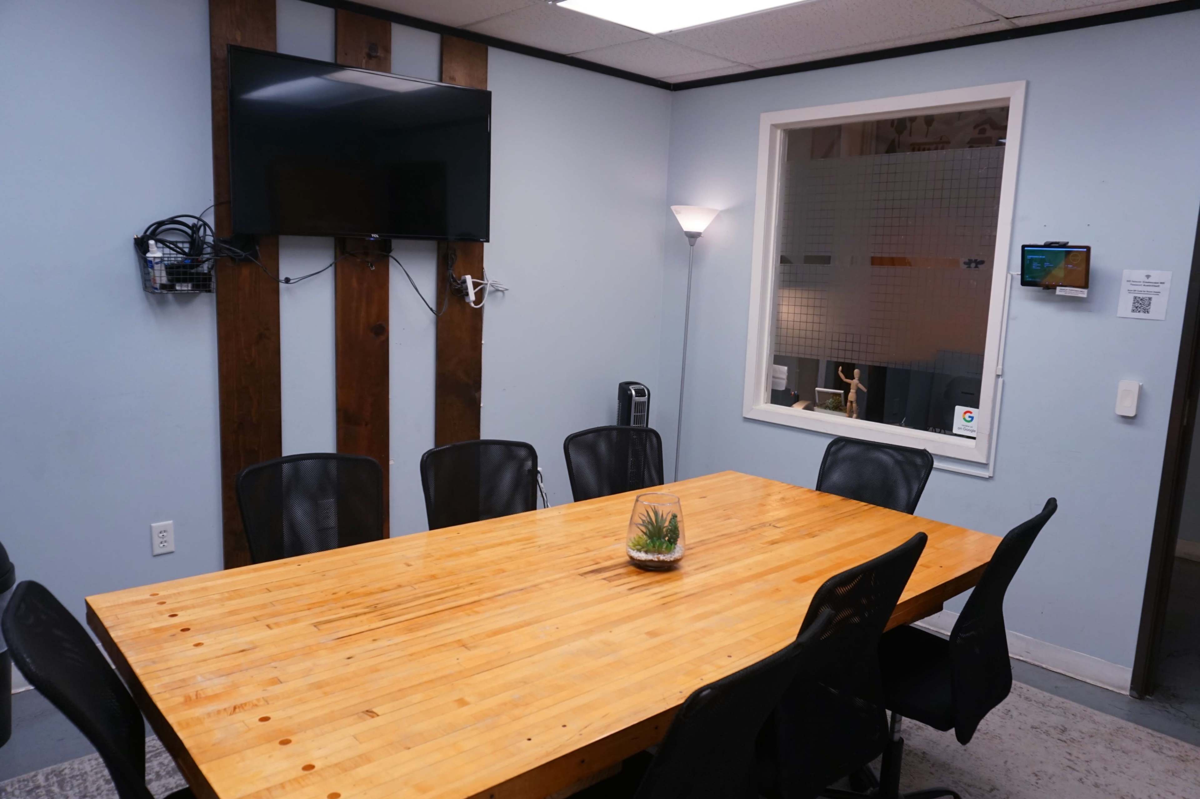 The image shows a conference room with a large wooden table surrounded by black chairs, a mounted television, and a window displaying a small indoor plant.